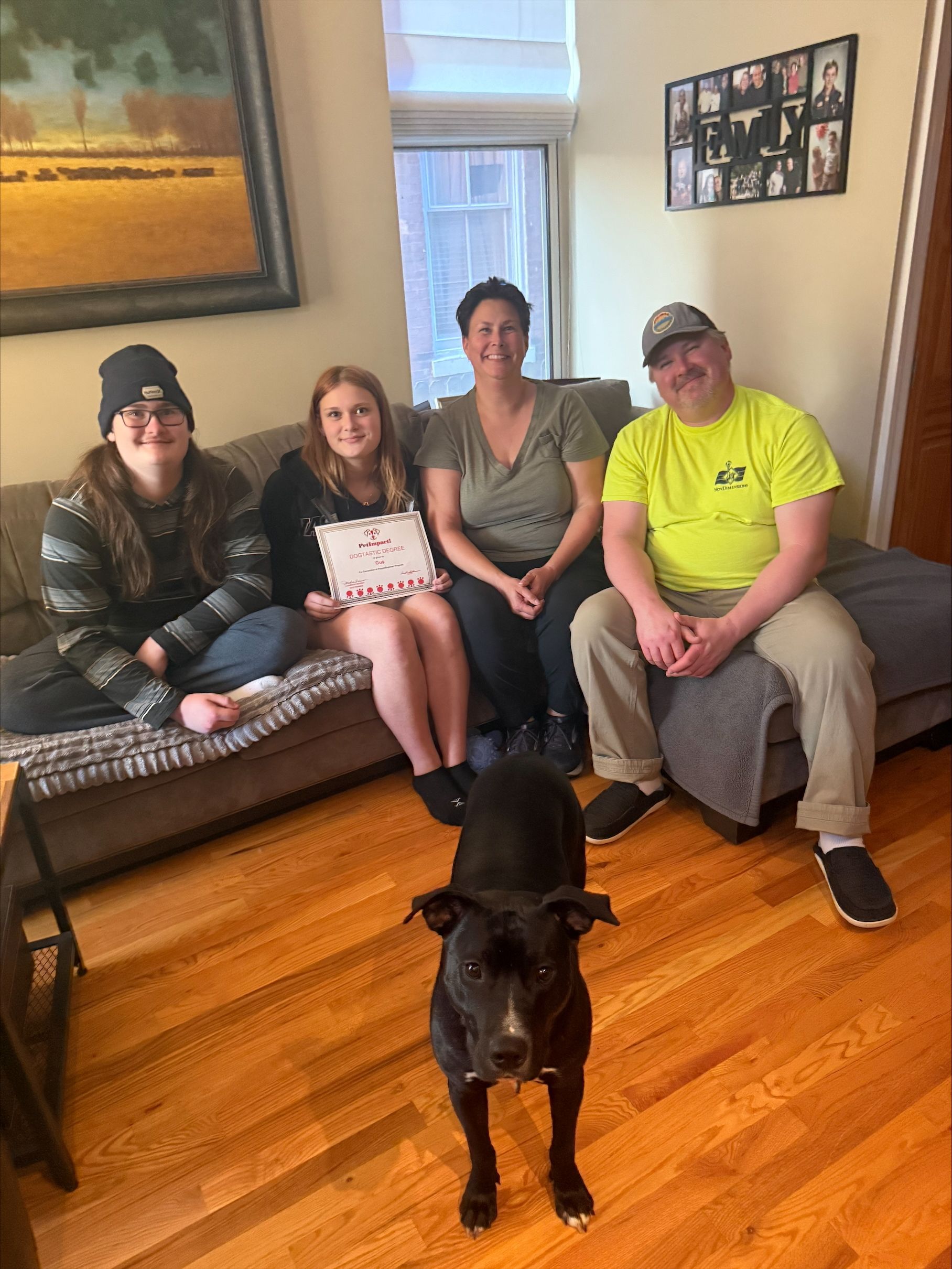 A family sits on a gray couch in a living room behind a black dog on a wood floor; they are smiling toward the camera.