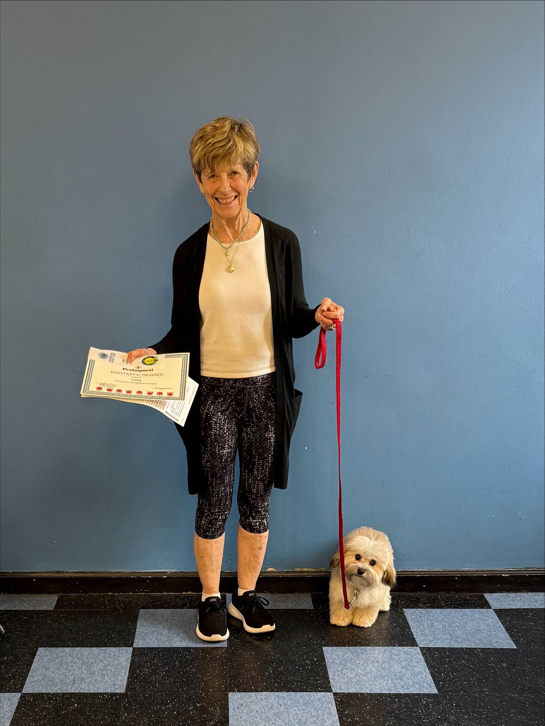 Woman with small dog, holding a certificate and leash. Standing in front of blue wall, patterned floor.