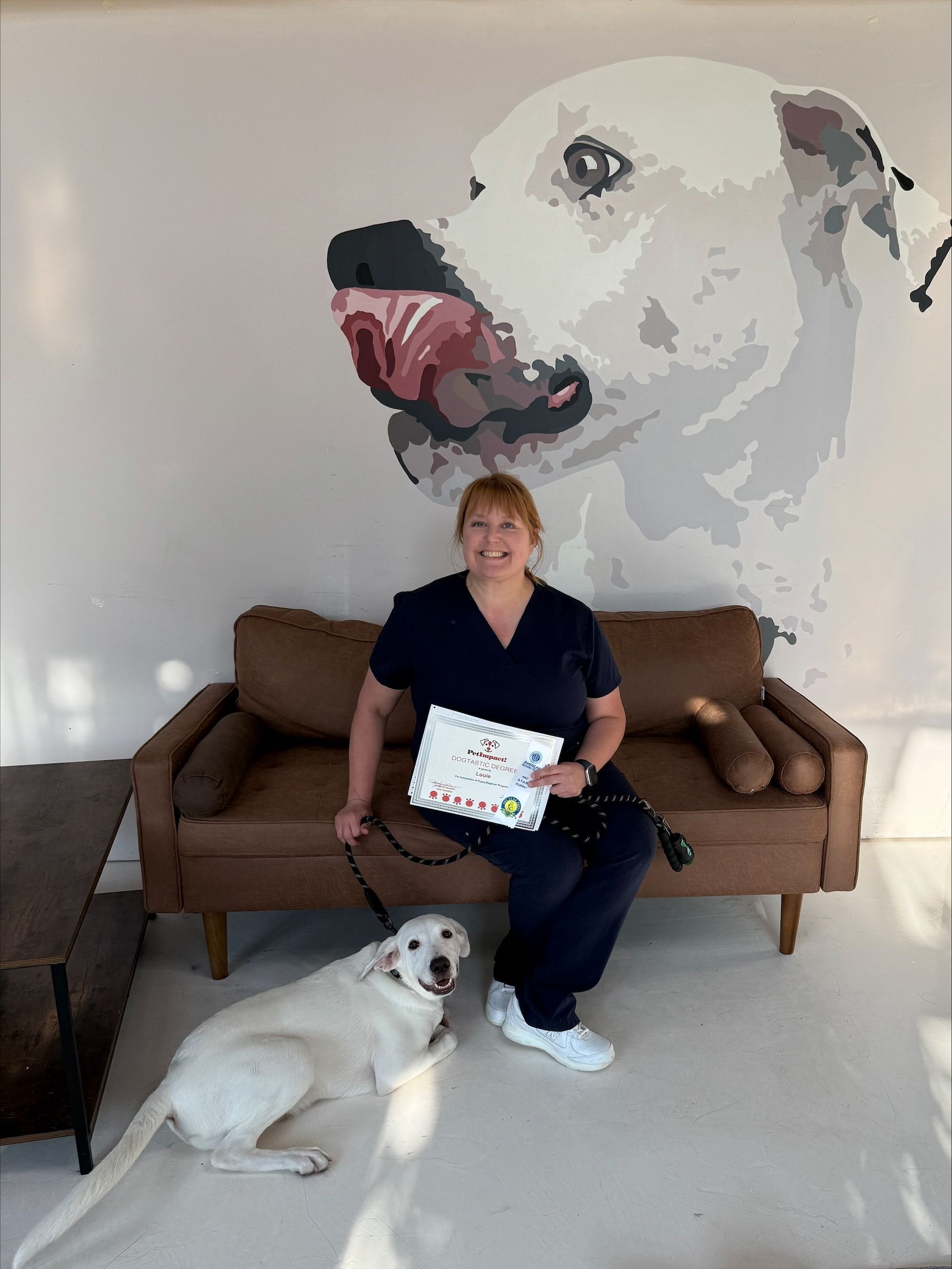 Woman in scrubs sits on couch with white dog and certificate, dog mural in the background.