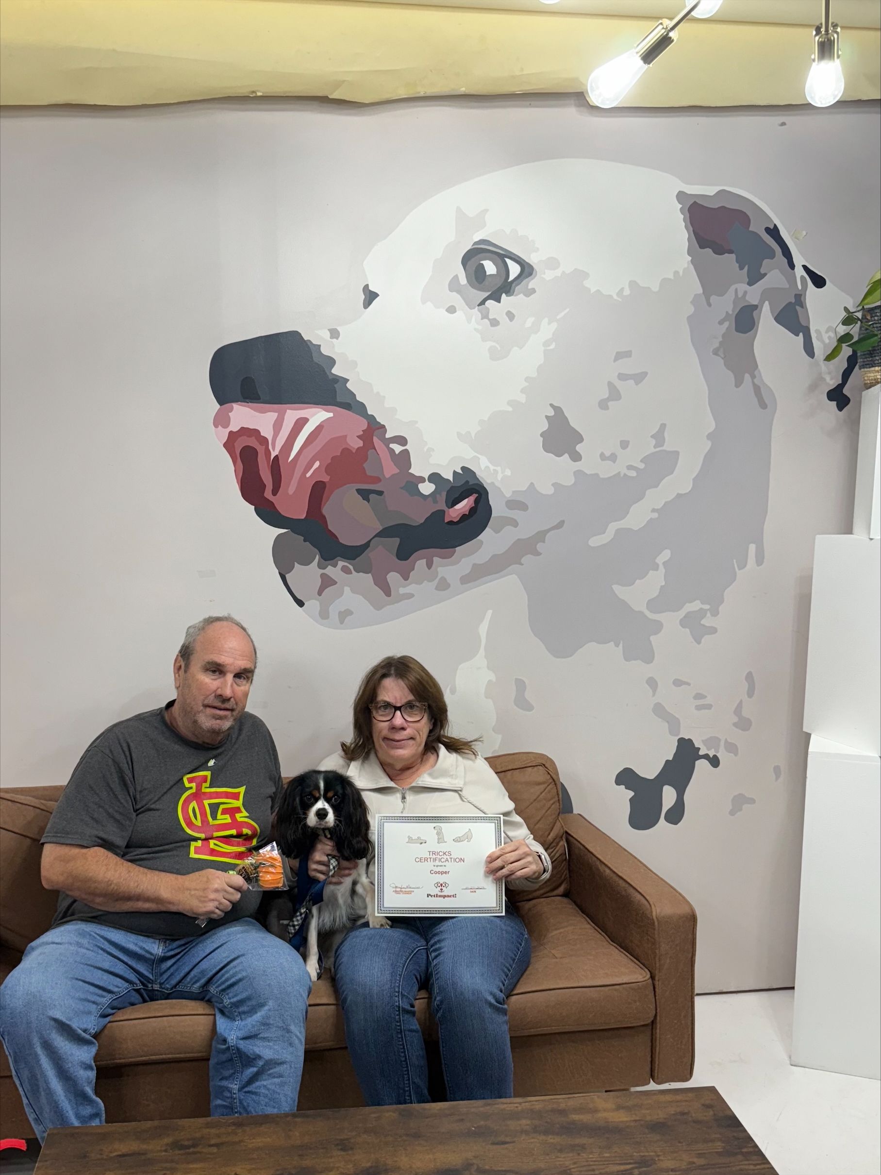 Couple with dog on couch posing in front of a giant dog mural. The woman holds a drawing of a dog.