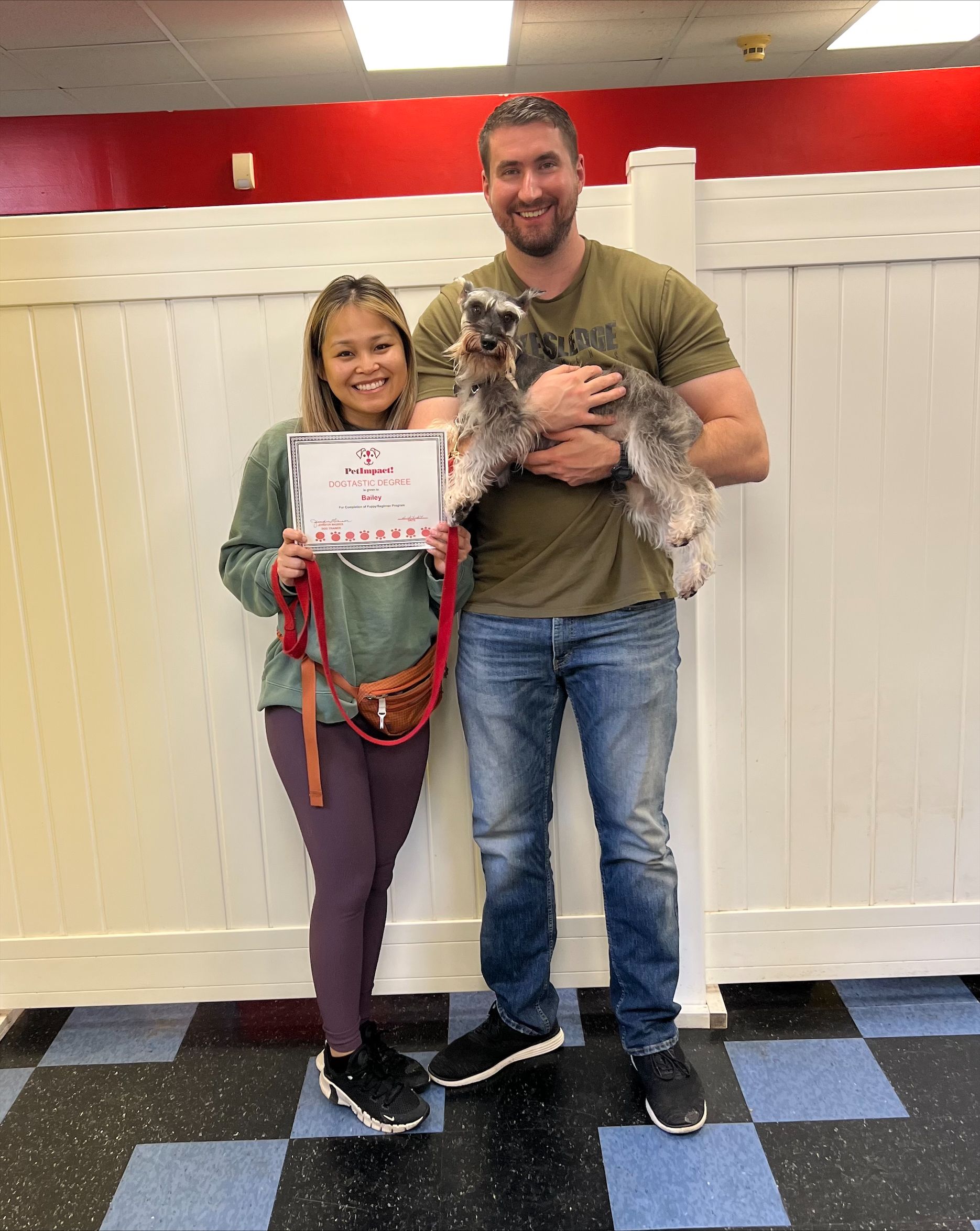 Woman holds certificate, man holds dog, both smiling in front of a white wall with red accents.
