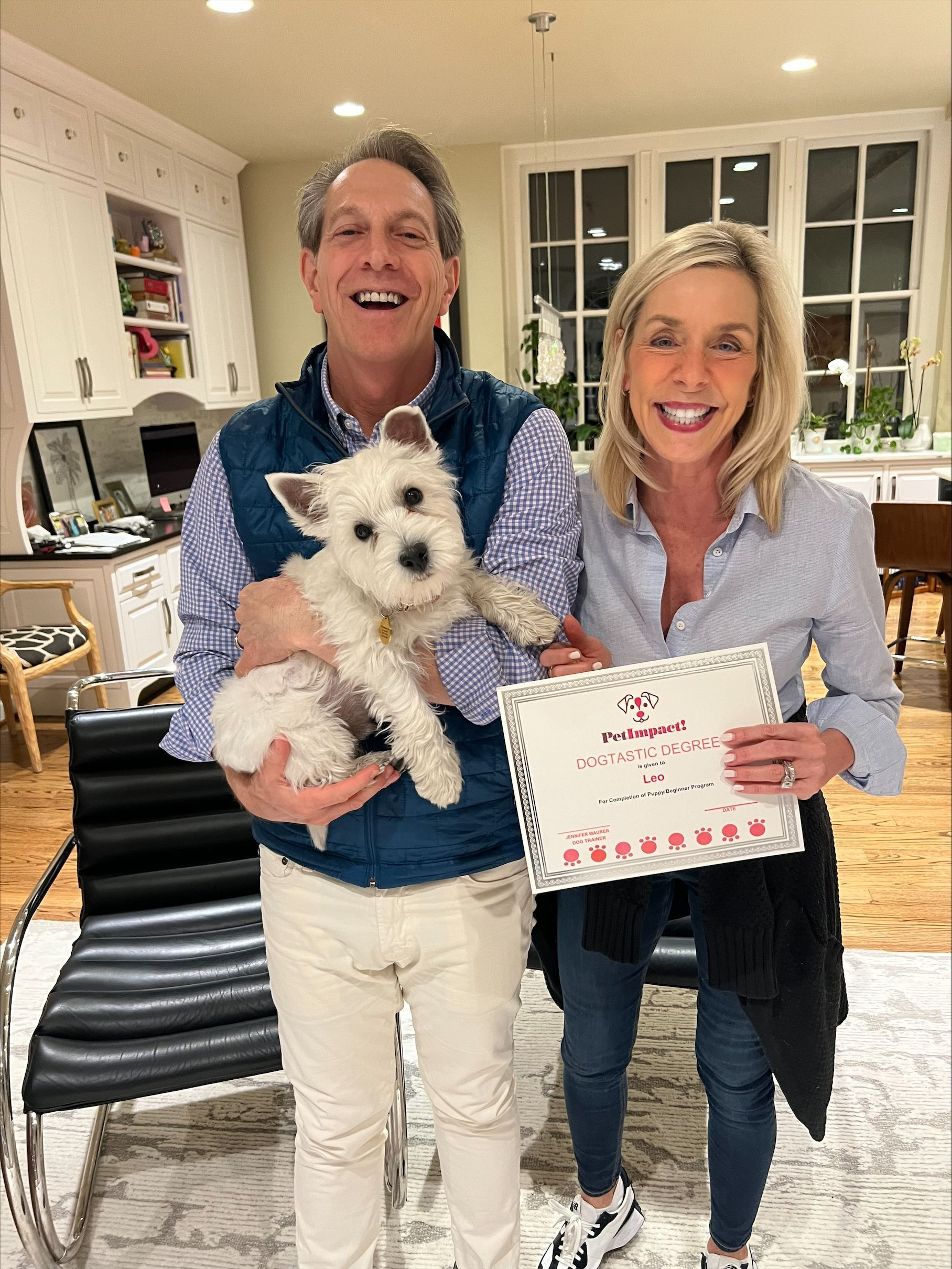 Man holds a white dog with a woman holding a certificate, both smiling. They are in a well-lit room.