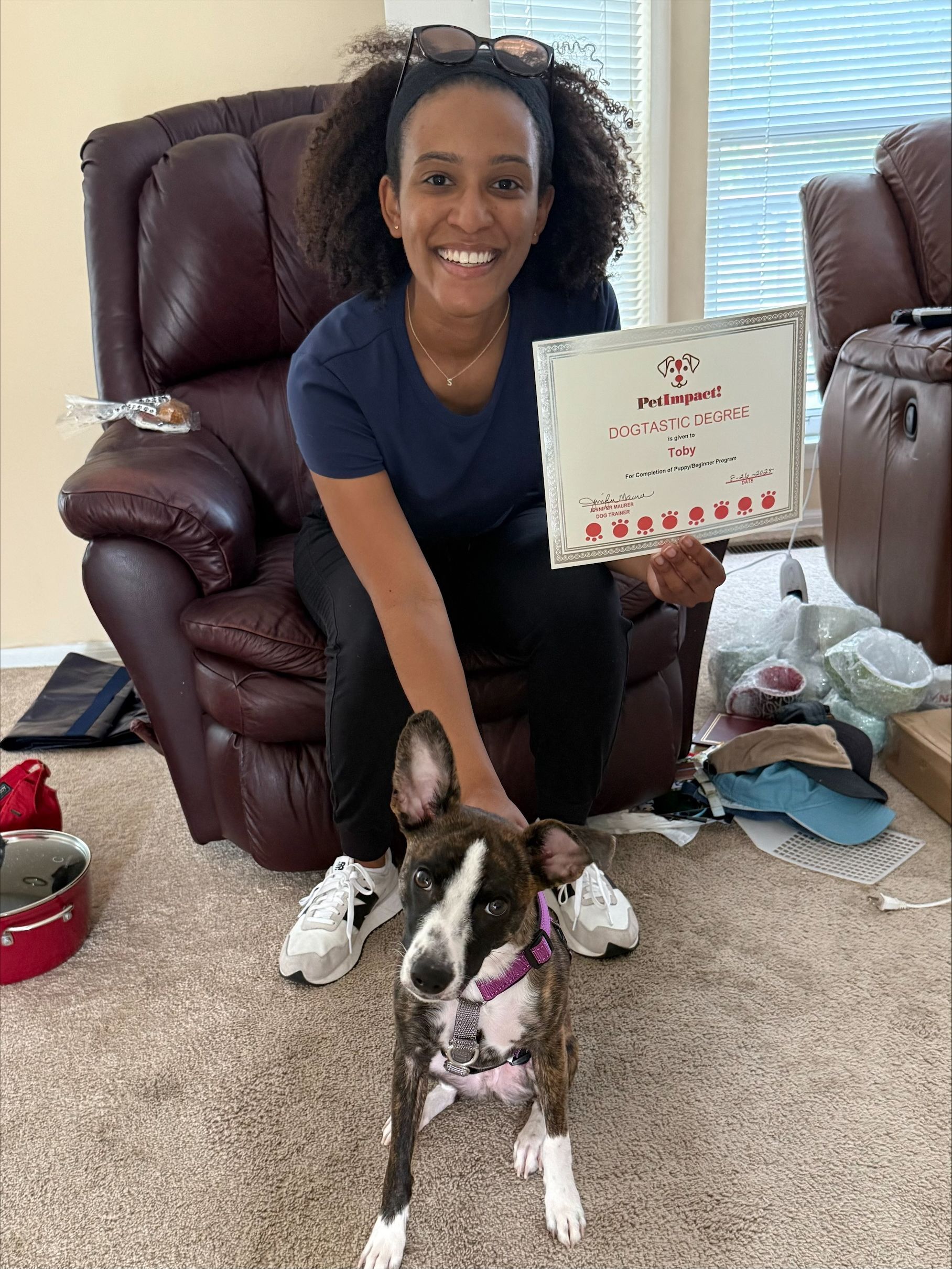 Woman smiling, petting a brindle dog; indoors, holding a sign.