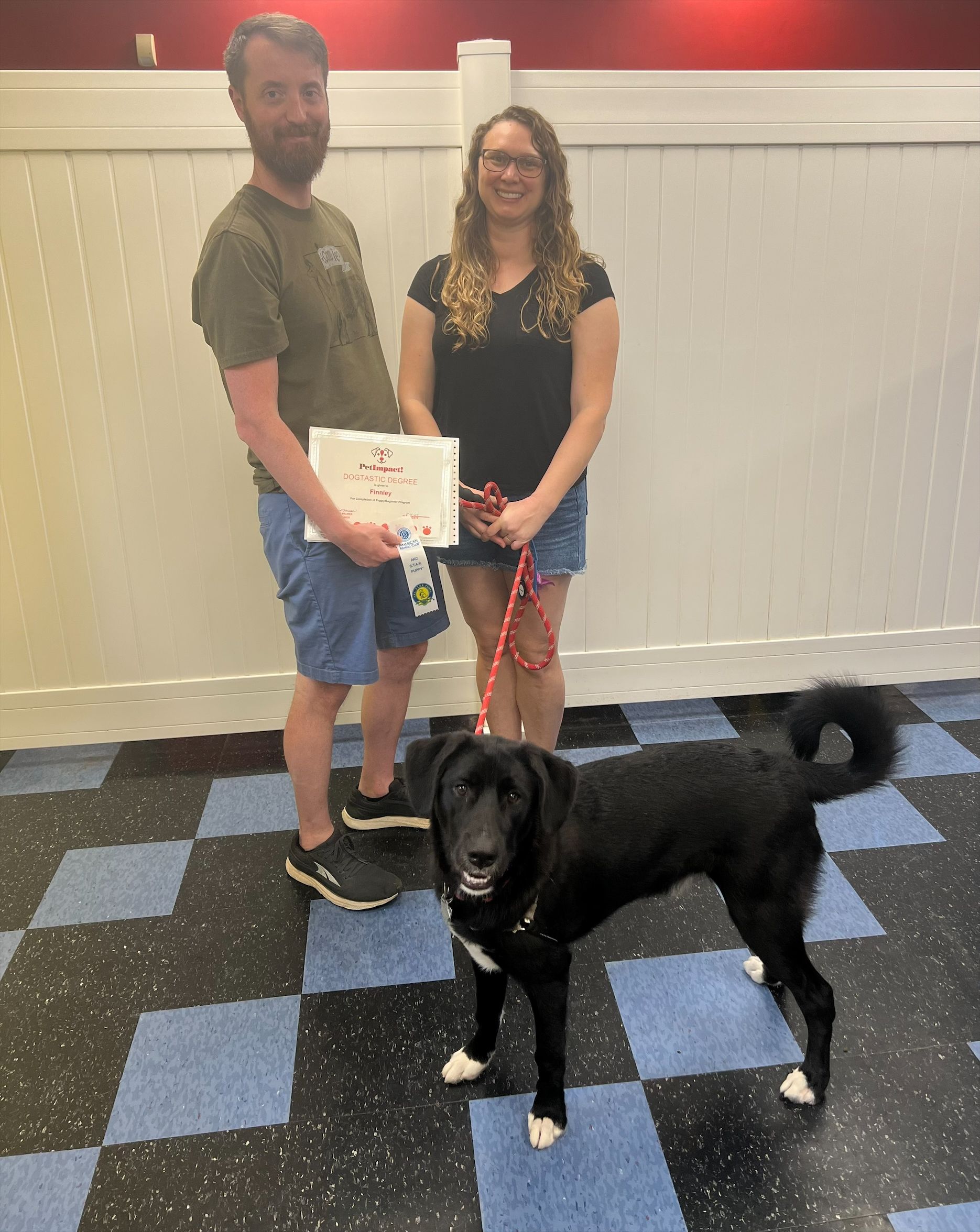 A man and a woman are standing next to a black dog on a leash.