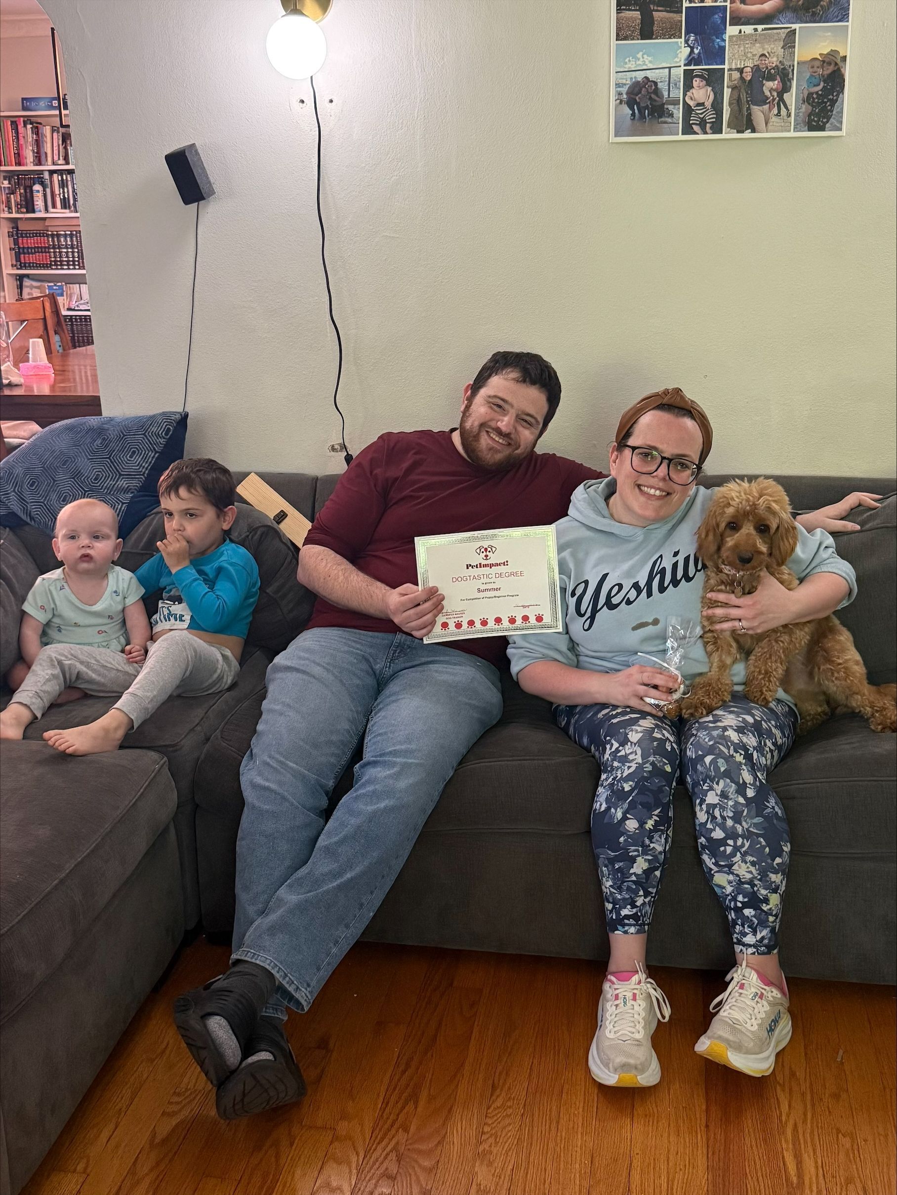 Family sitting on a couch with two children and a dog, smiling indoors; one person holds a sign.