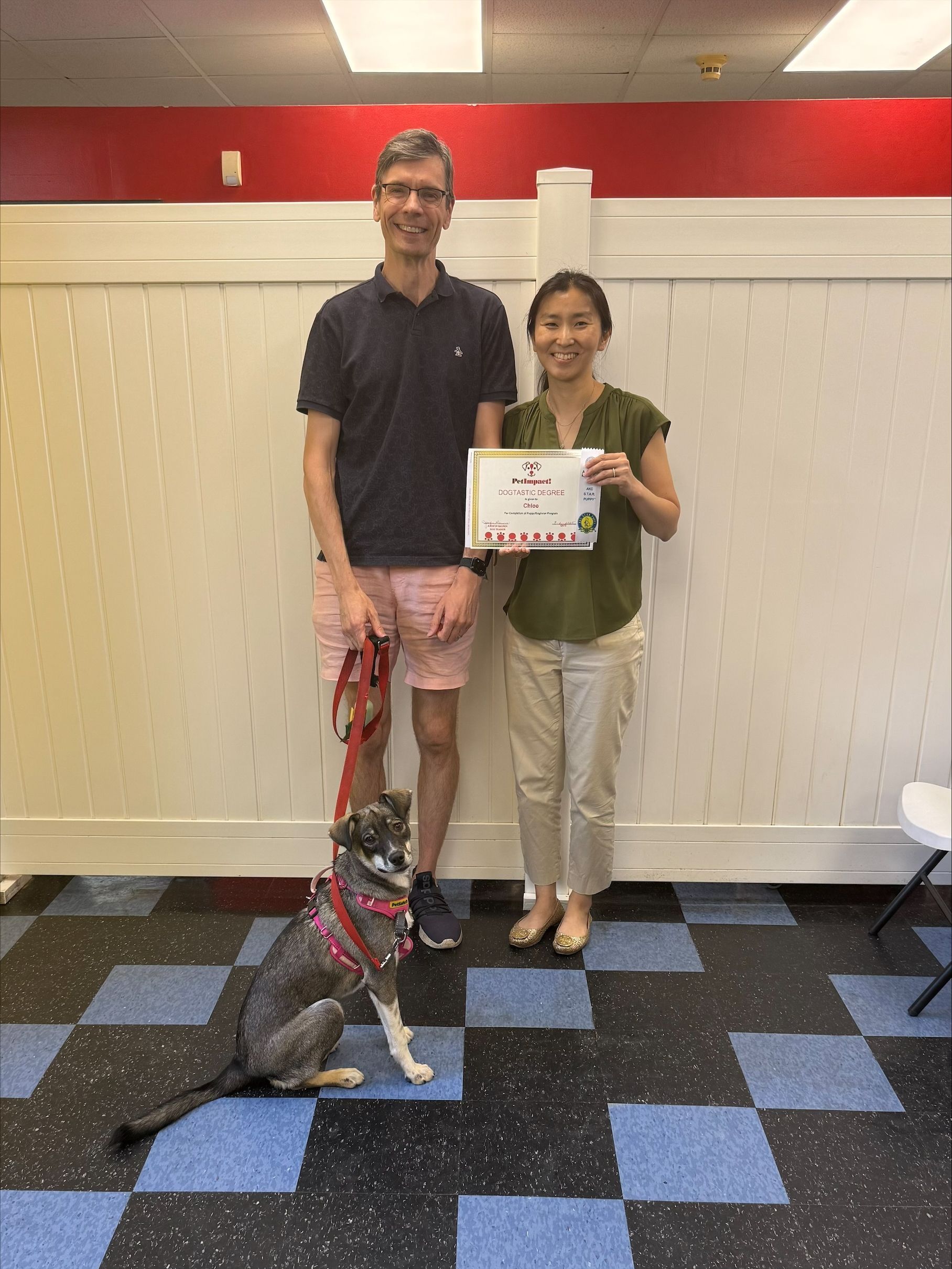 Man and woman with dog holding a certificate. They are in a room with a blue and black checkered floor, red wall and white paneling.