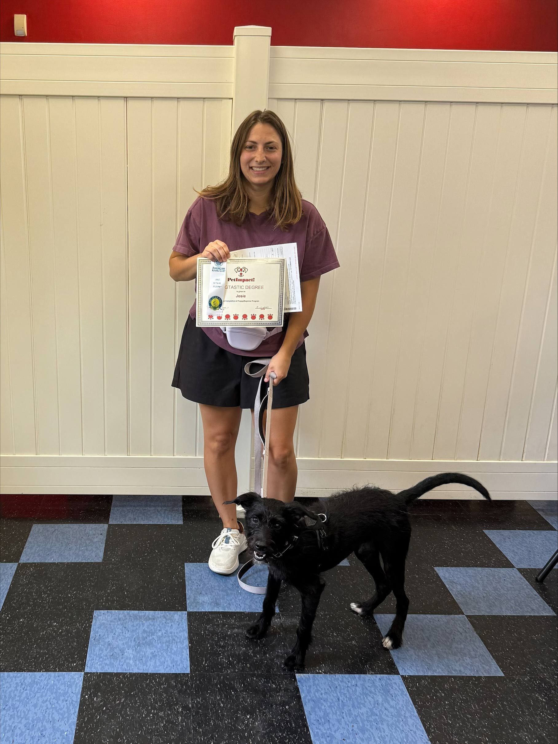 Woman holding certificate, smiles next to black dog with leash indoors; red wall, white panels, and patterned floor.