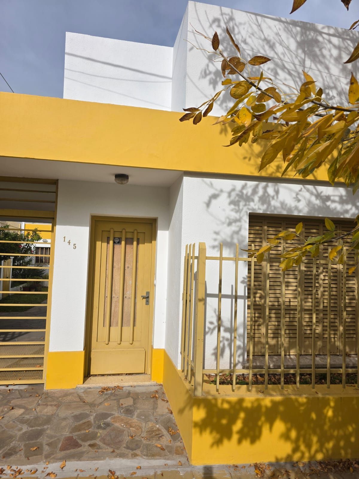 Entrada a una pequeña casa con paredes amarillas y blancas, puerta de madera y verja metálica, bañada por la luz del sol.