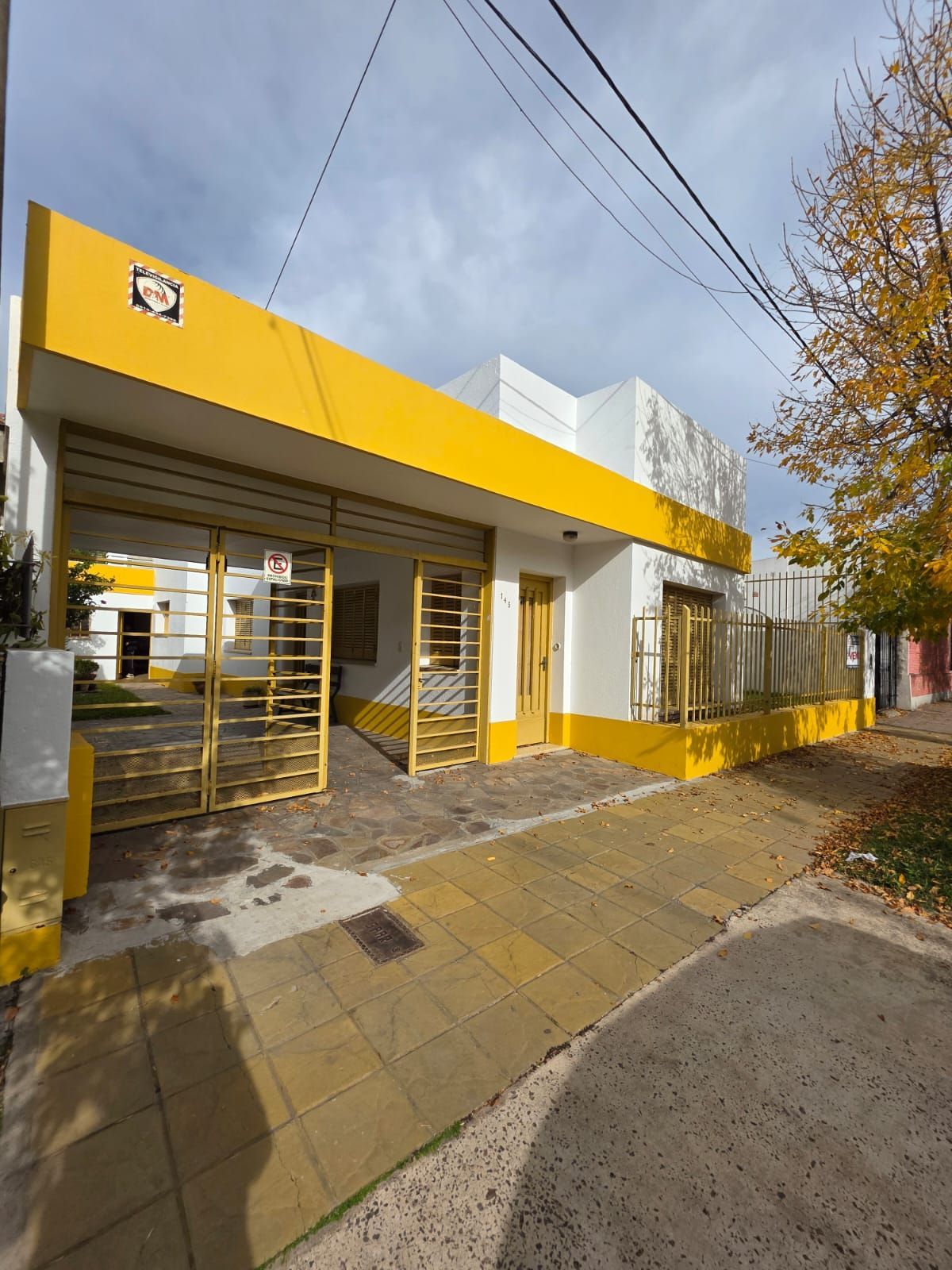 Edificio amarillo y blanco con entrada cerrada y acera bajo árboles otoñales.