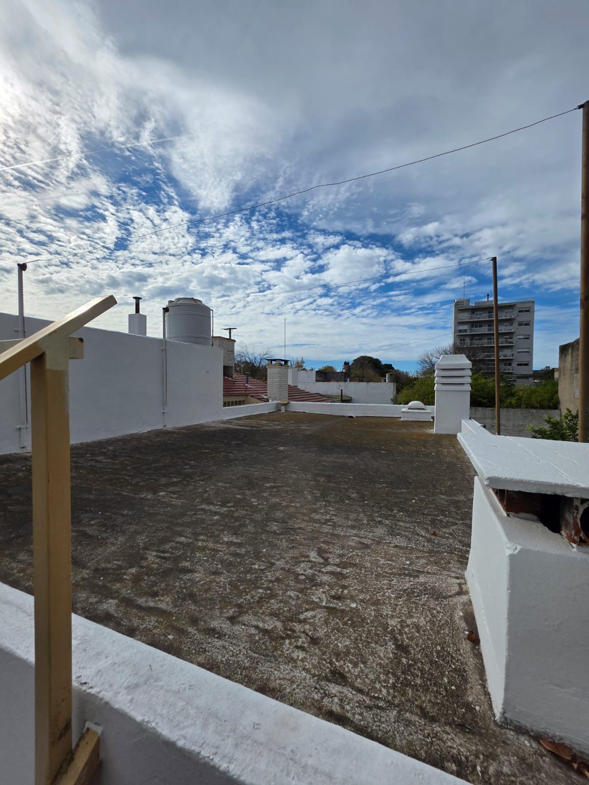 Terraza en la azotea con paredes blancas, suelo de grava y un cielo nublado sobre los edificios cercanos.
