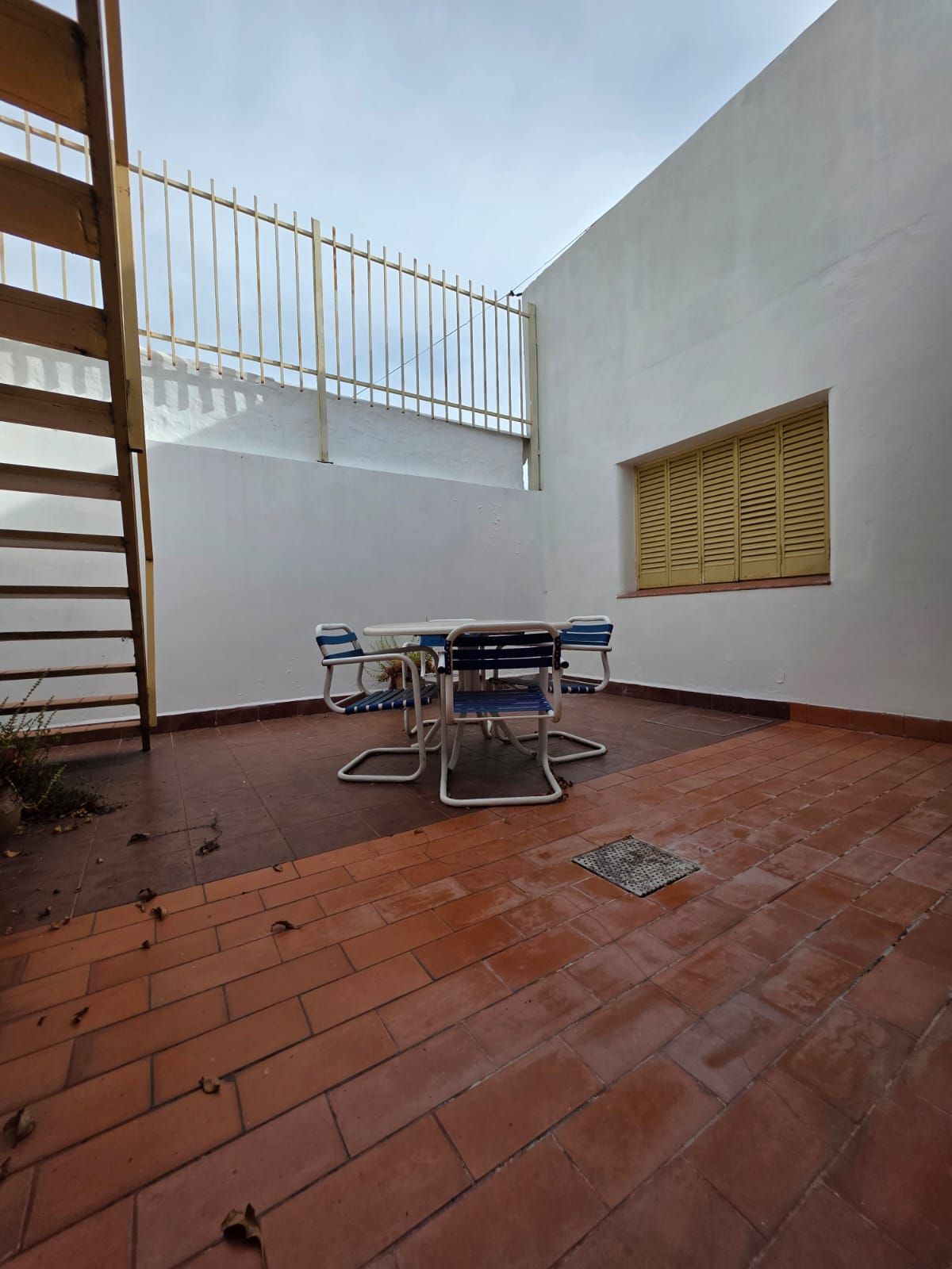 Pequeño patio embaldosado con una mesa redonda y sillas, paredes blancas y una escalera a la izquierda.