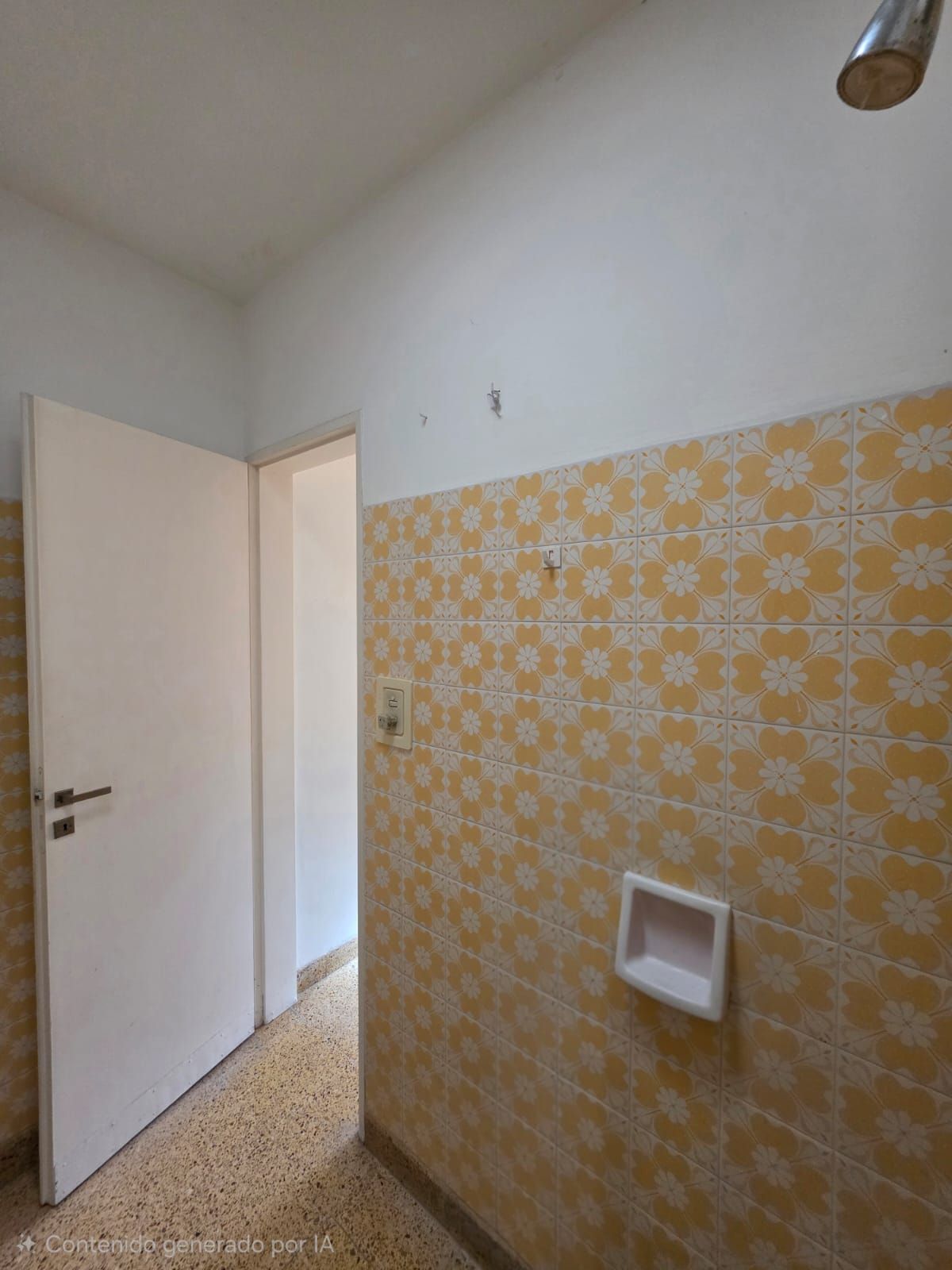 Habitación vacía con la puerta abierta, azulejos amarillos con estampado en la pared y una pequeña lámpara de pared.