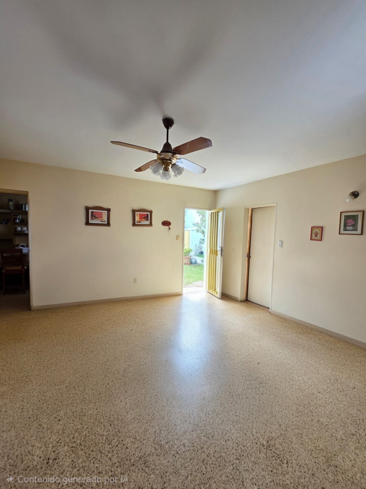 Sala de estar vacía de color beige con ventilador de techo, piso de baldosas y puerta abierta a un luminoso exterior.