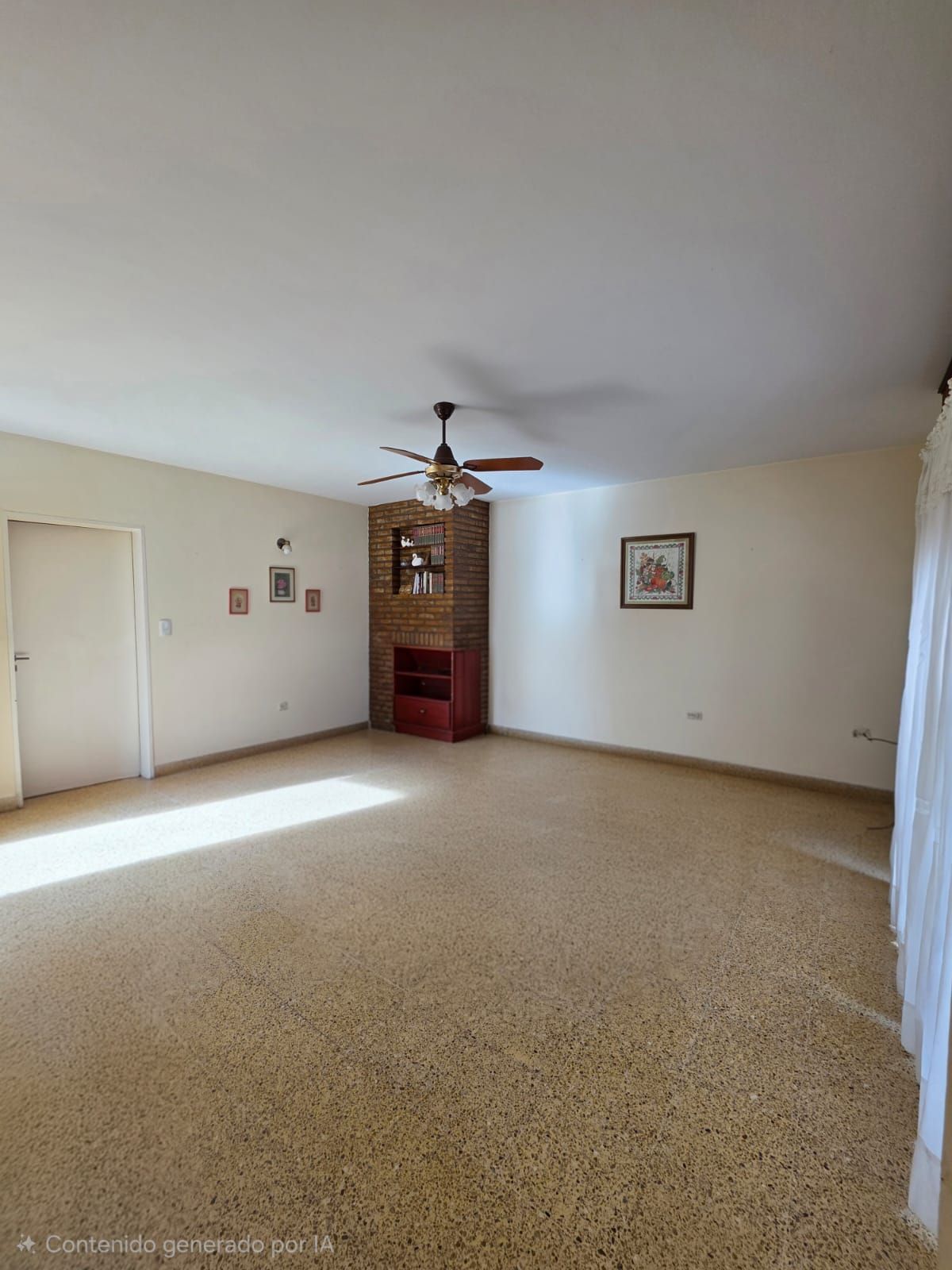 Sala de estar vacía de color beige con ventilador de techo, chimenea de ladrillo y luz solar sobre la alfombra.