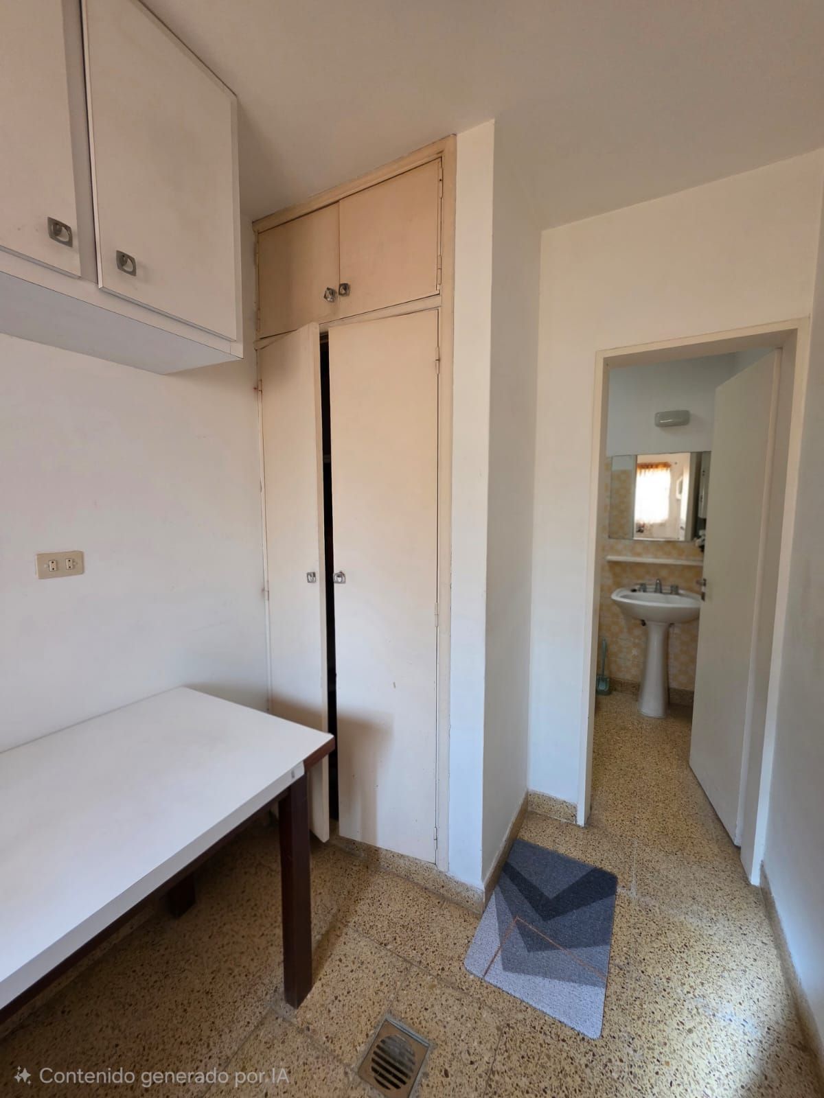 Pequeña cocina americana con armarios blancos, mesa y una puerta que da al baño.