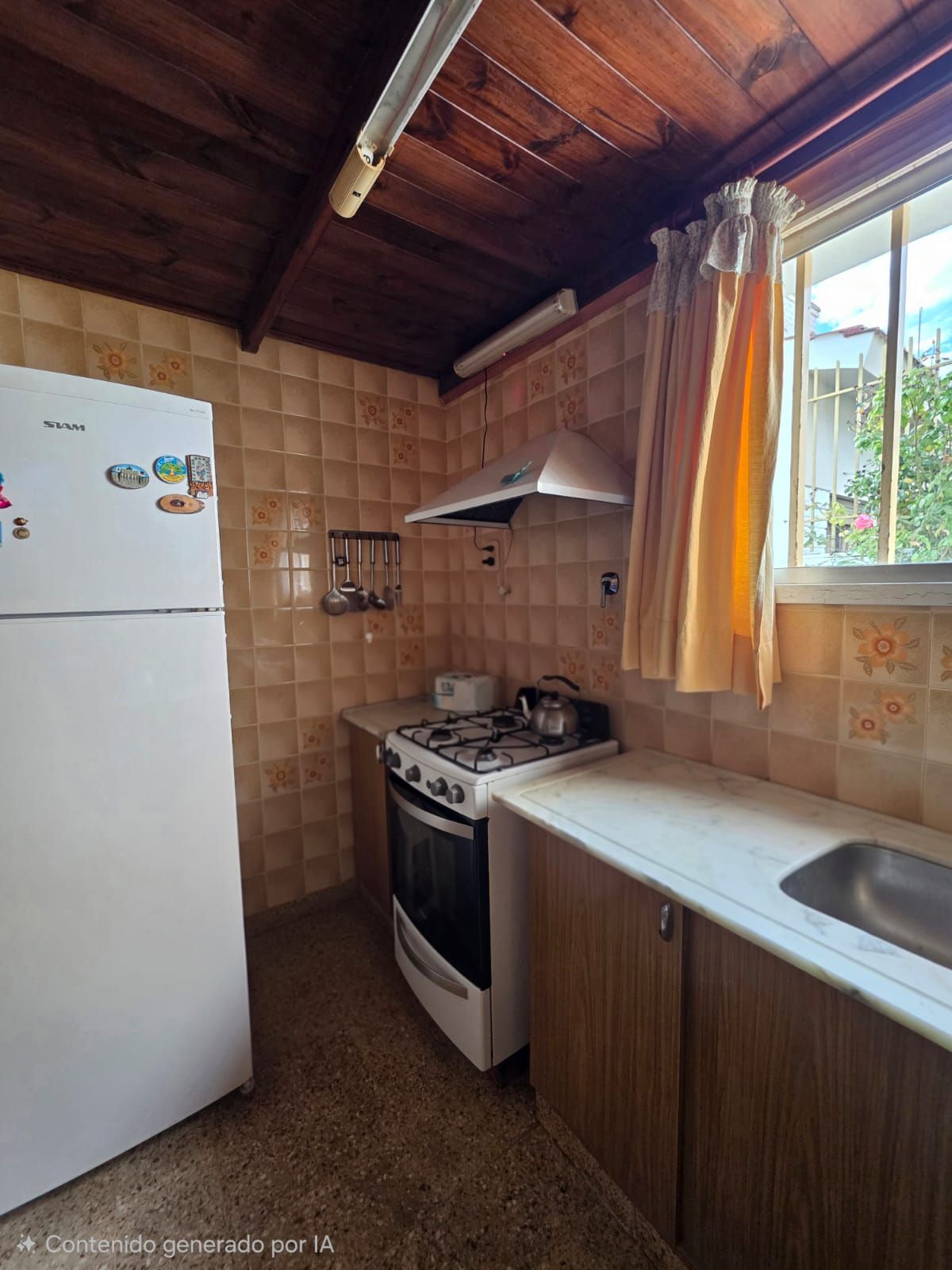 Cocina rústica compacta con nevera blanca, fogones, fregadero, techo de madera y una ventana con cortinas naranjas.