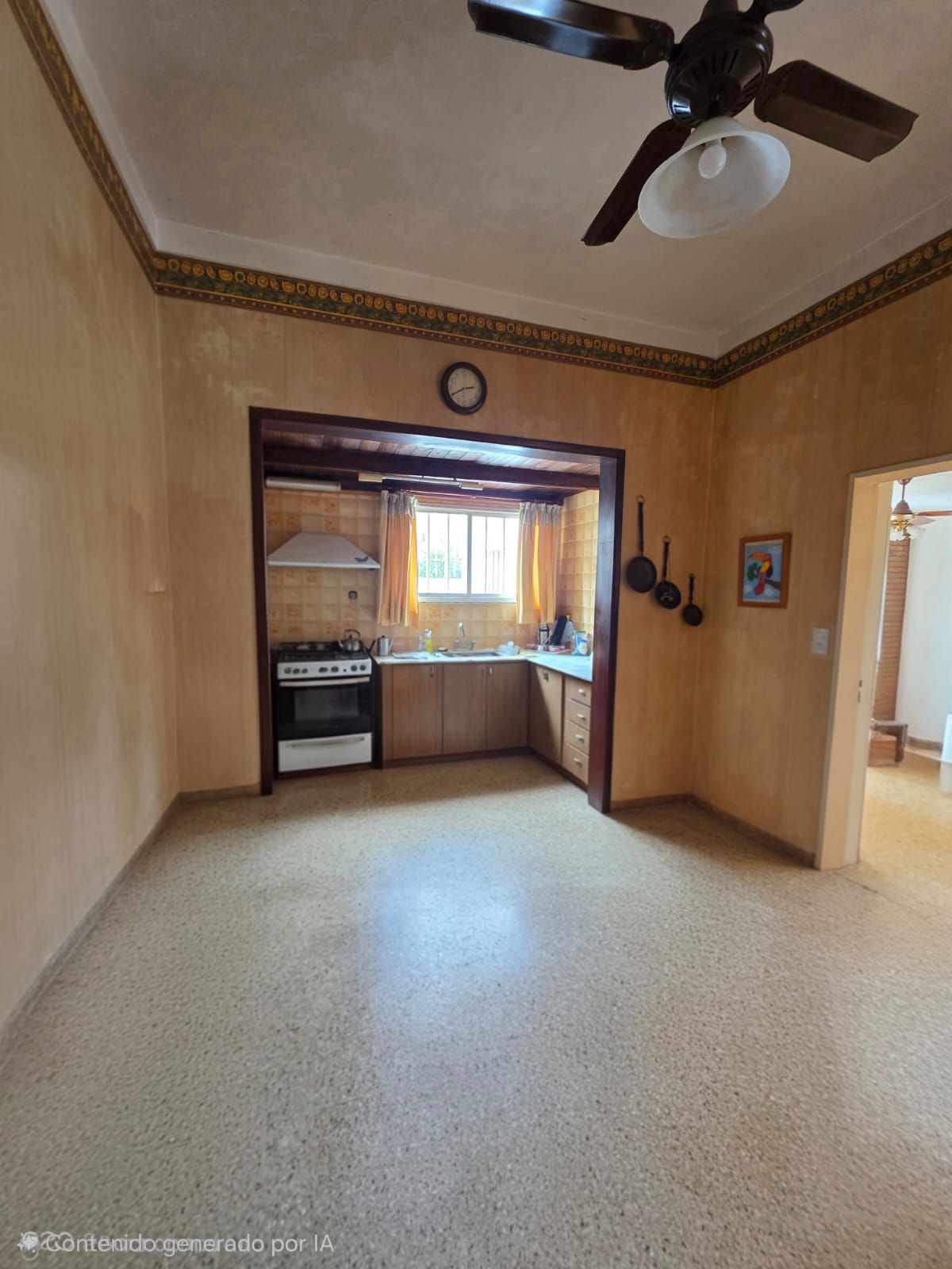 Comedor vacío de color beige que da a una cocina con armarios de madera, estufa, fregadero y ventilador de techo.