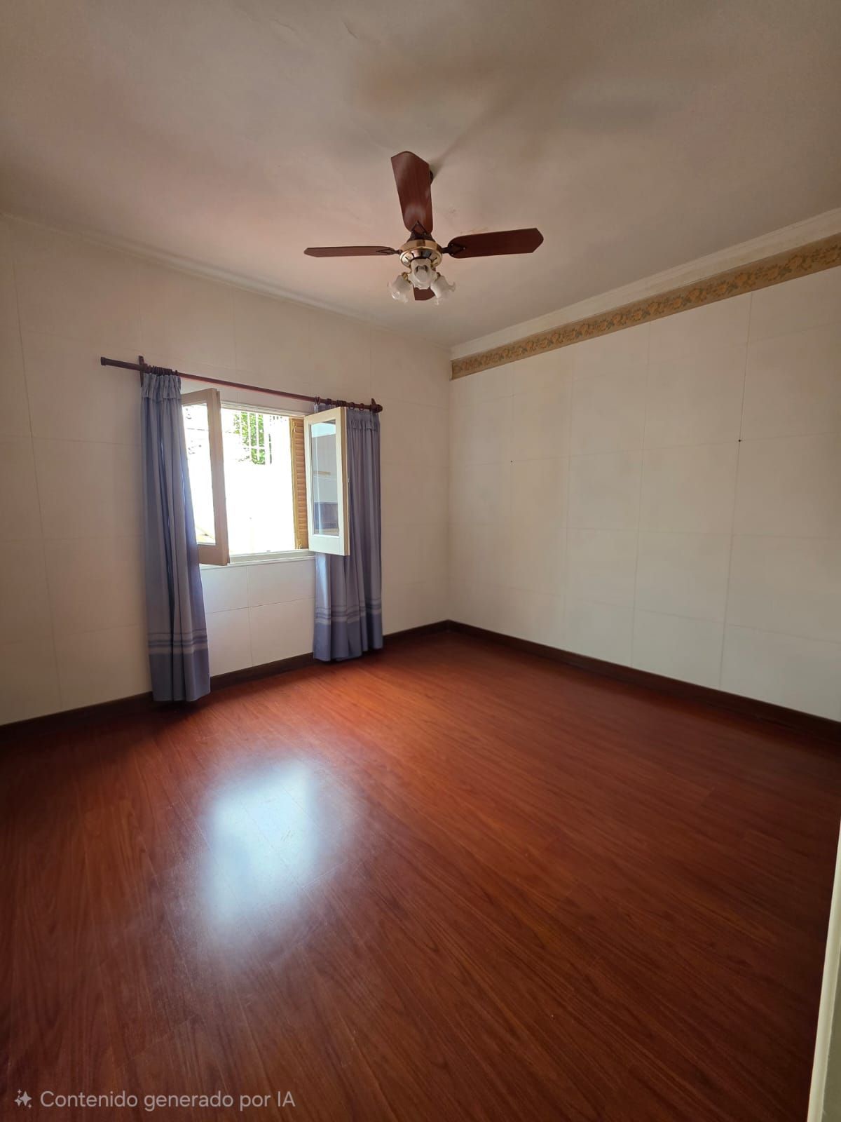 Habitación vacía con suelo de madera, paredes blancas, una ventana con cortinas azules y un ventilador de techo.