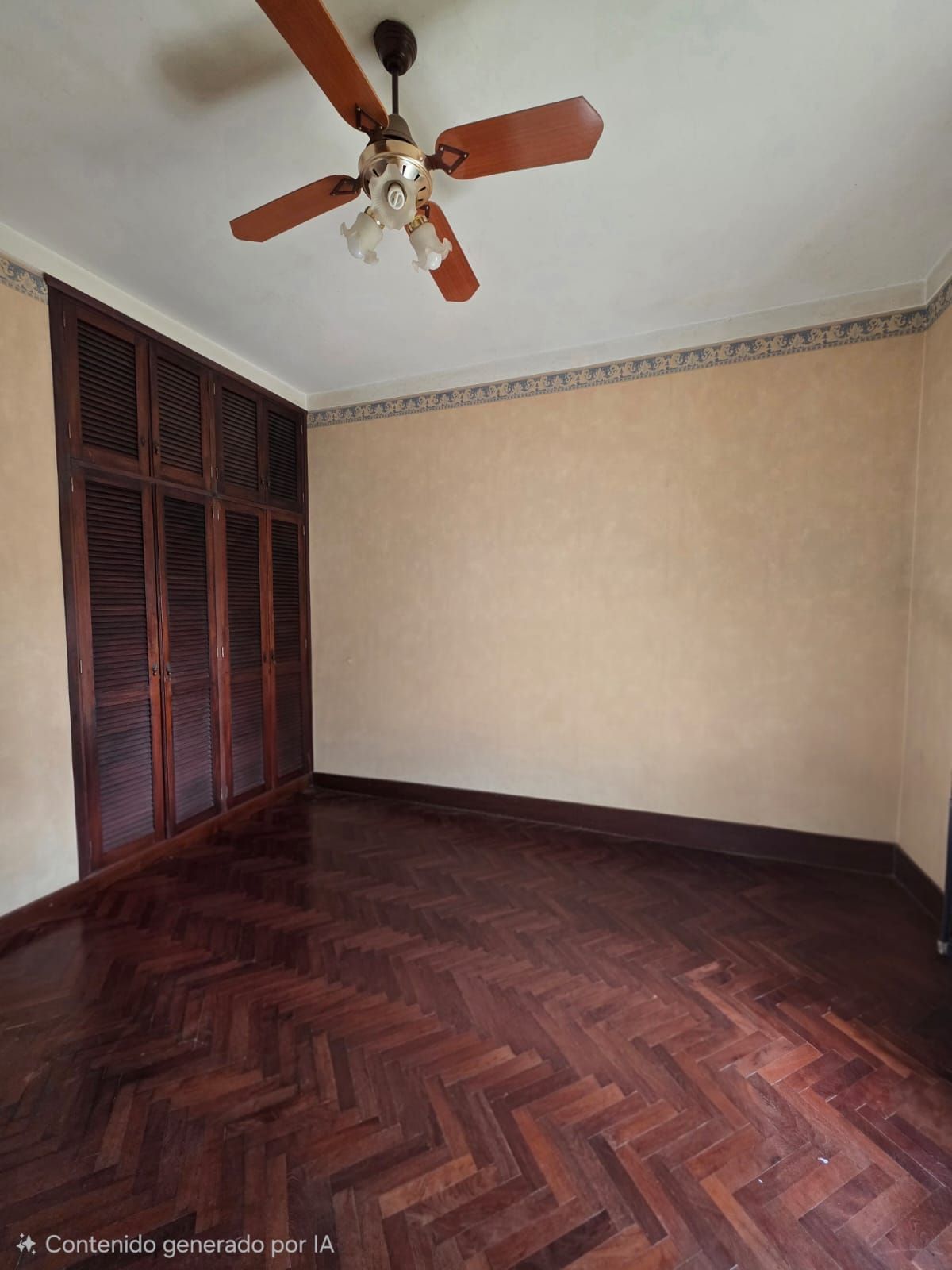 Habitación vacía con suelo de madera oscura en espiga, paredes beige, armario de madera y ventilador de techo.