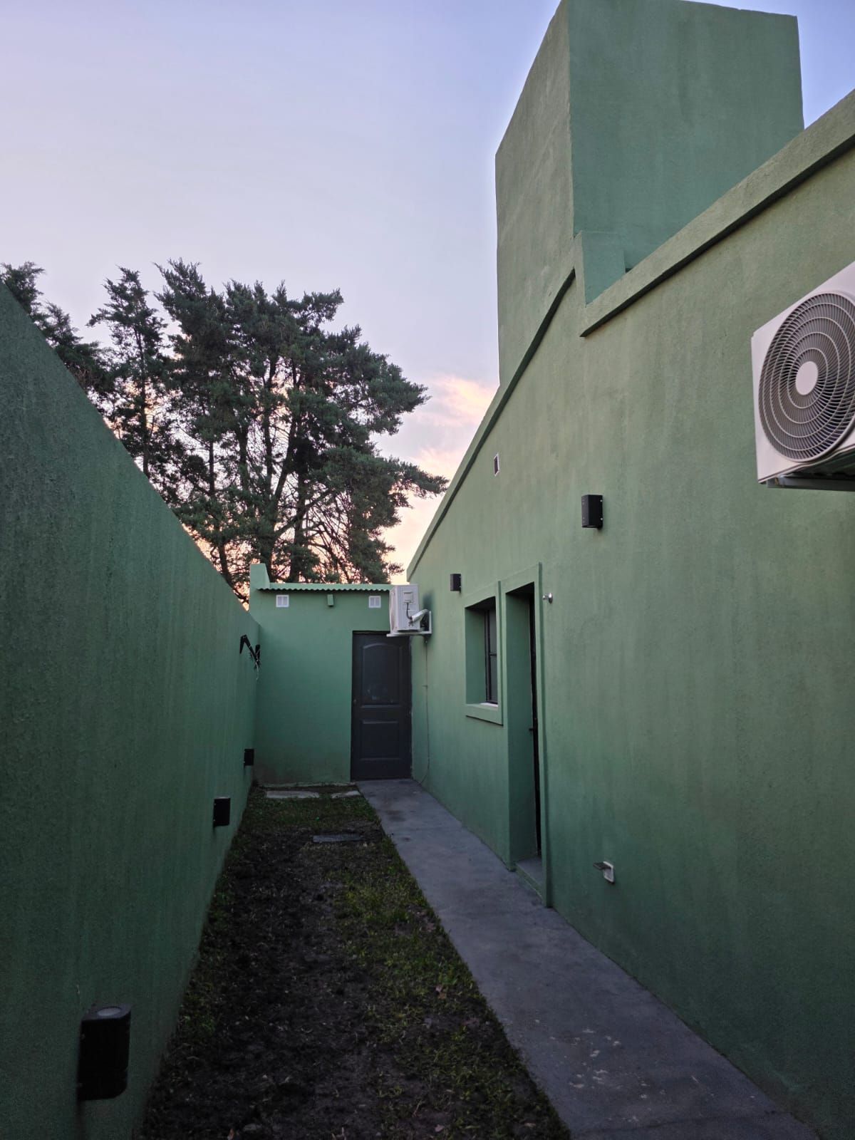 Estrecho pasillo verde en un edificio al anochecer, con una puerta lateral y una unidad de aire acondicionado.