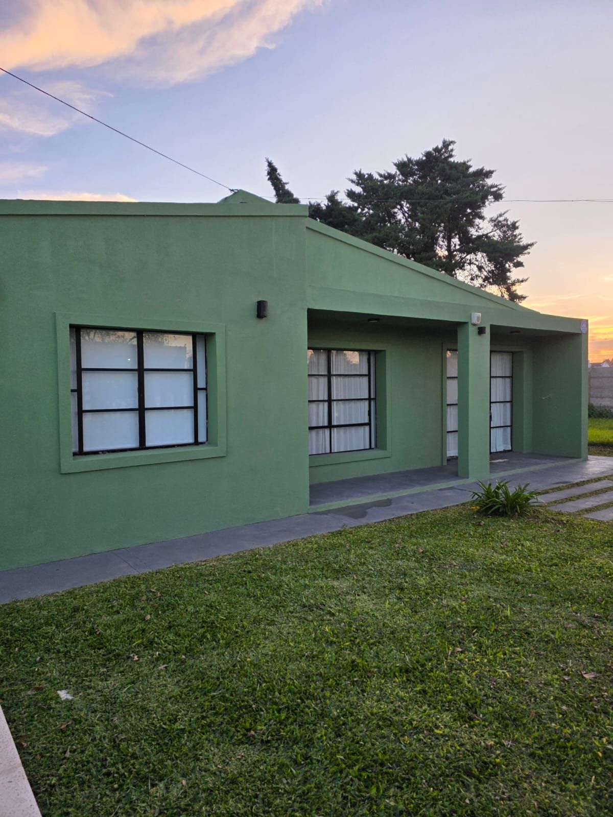Casa verde de una sola planta con ventanas y un pequeño porche, vista junto a un jardín cubierto de césped al atardecer.