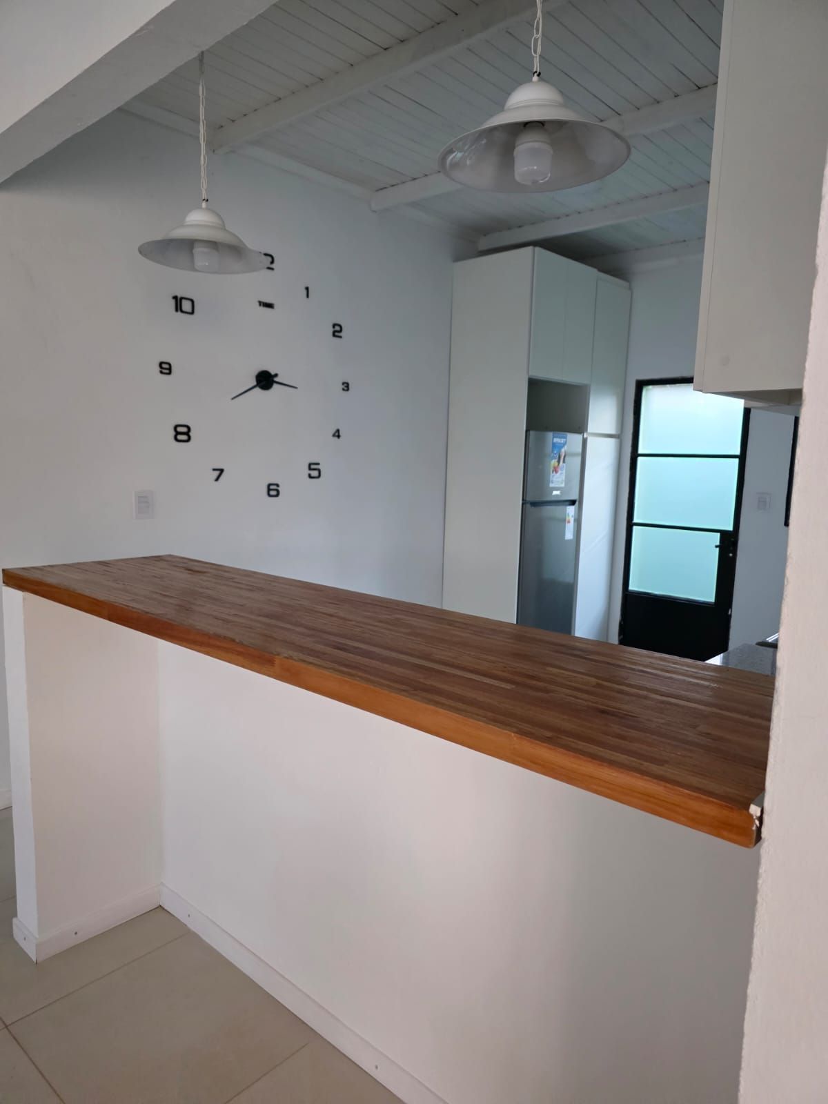 Interior de cocina minimalista con barra de desayuno de madera, reloj de pared, ventiladores de techo y refrigerador.