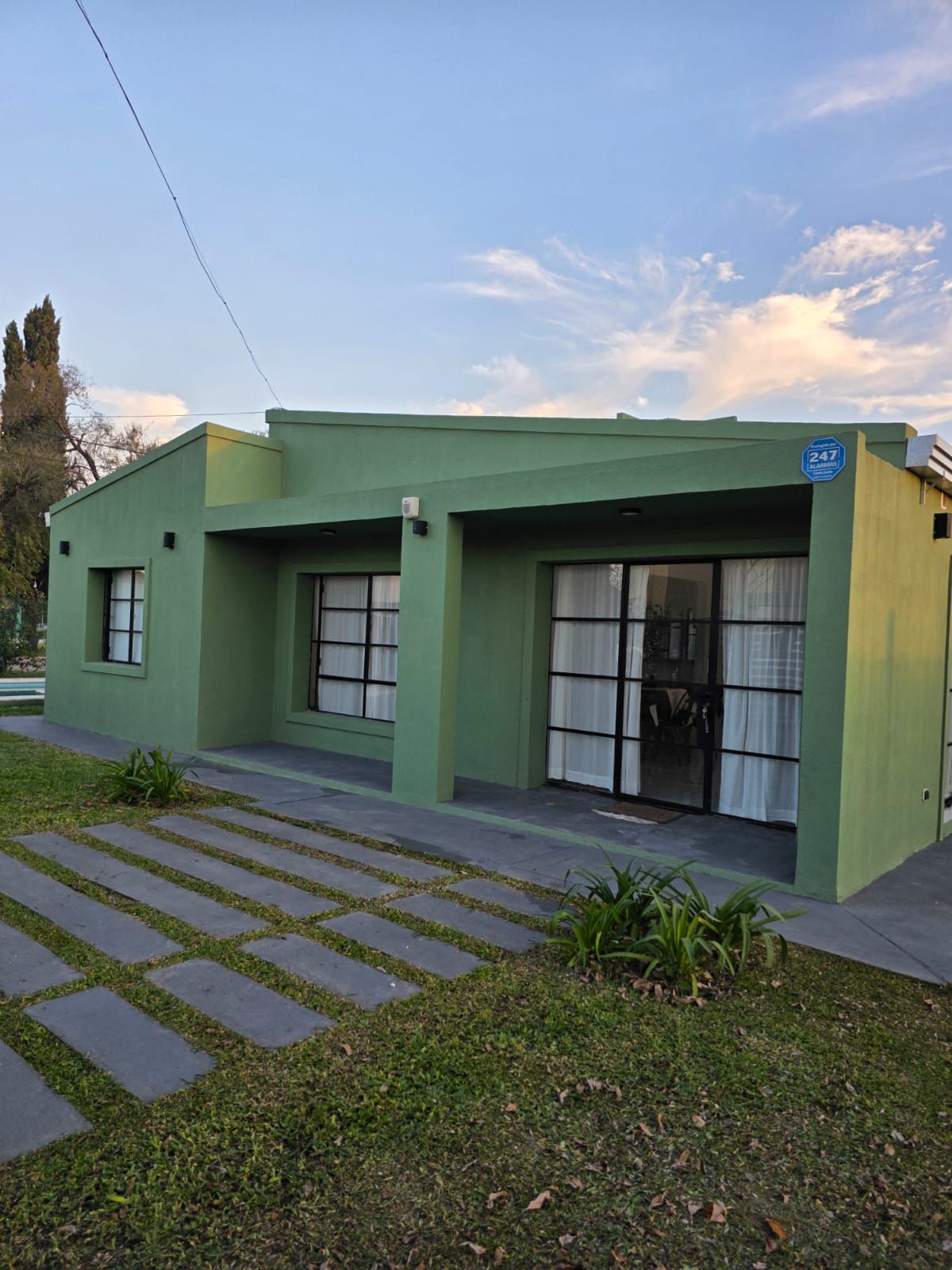 Casa verde de una sola planta con grandes ventanales, sendero a rayas y pequeño patio delantero al atardecer.