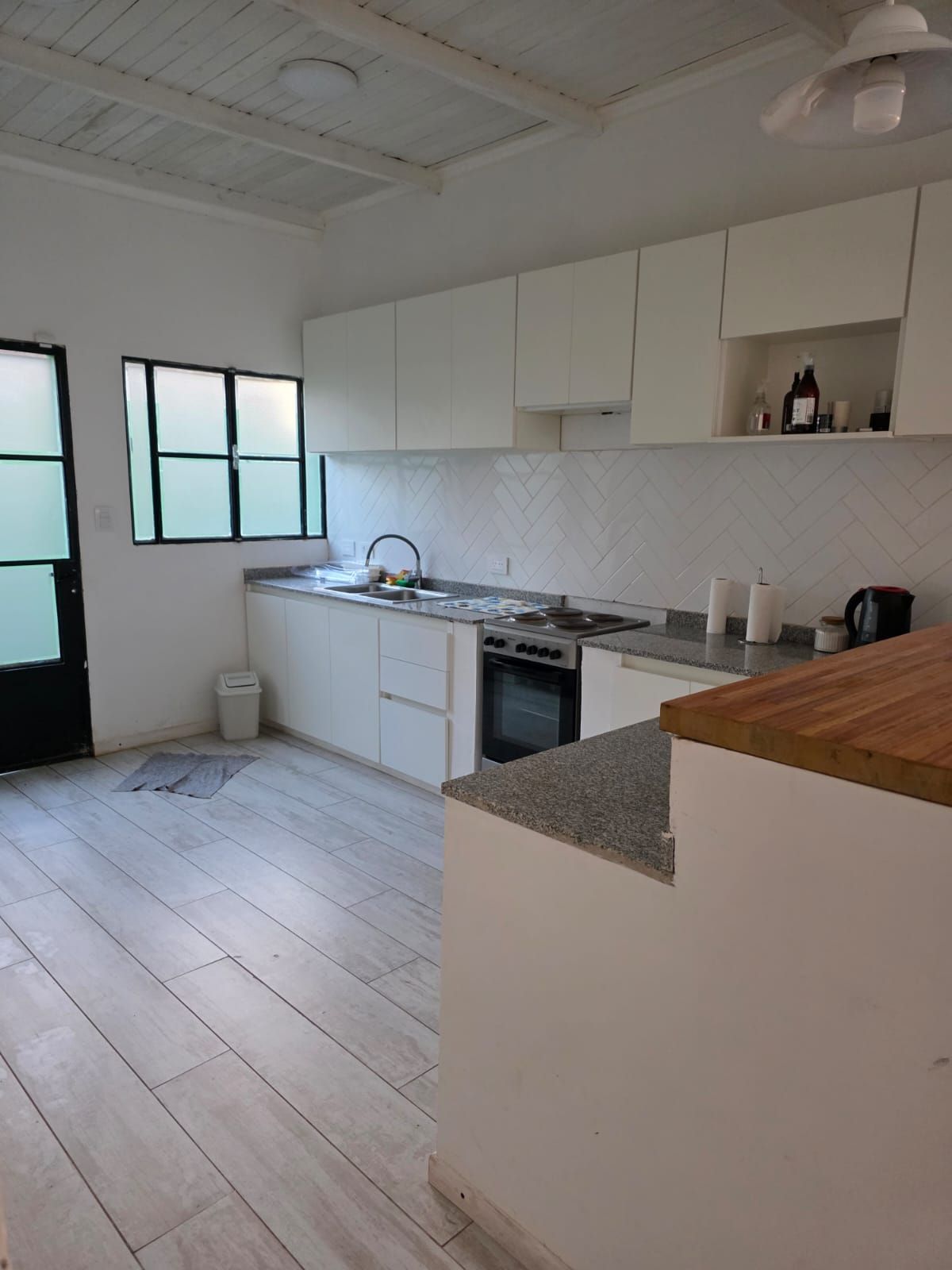 Cocina luminosa y sin amueblar, con armarios blancos, encimeras de madera, salpicadero de azulejos y puerta con ventana.