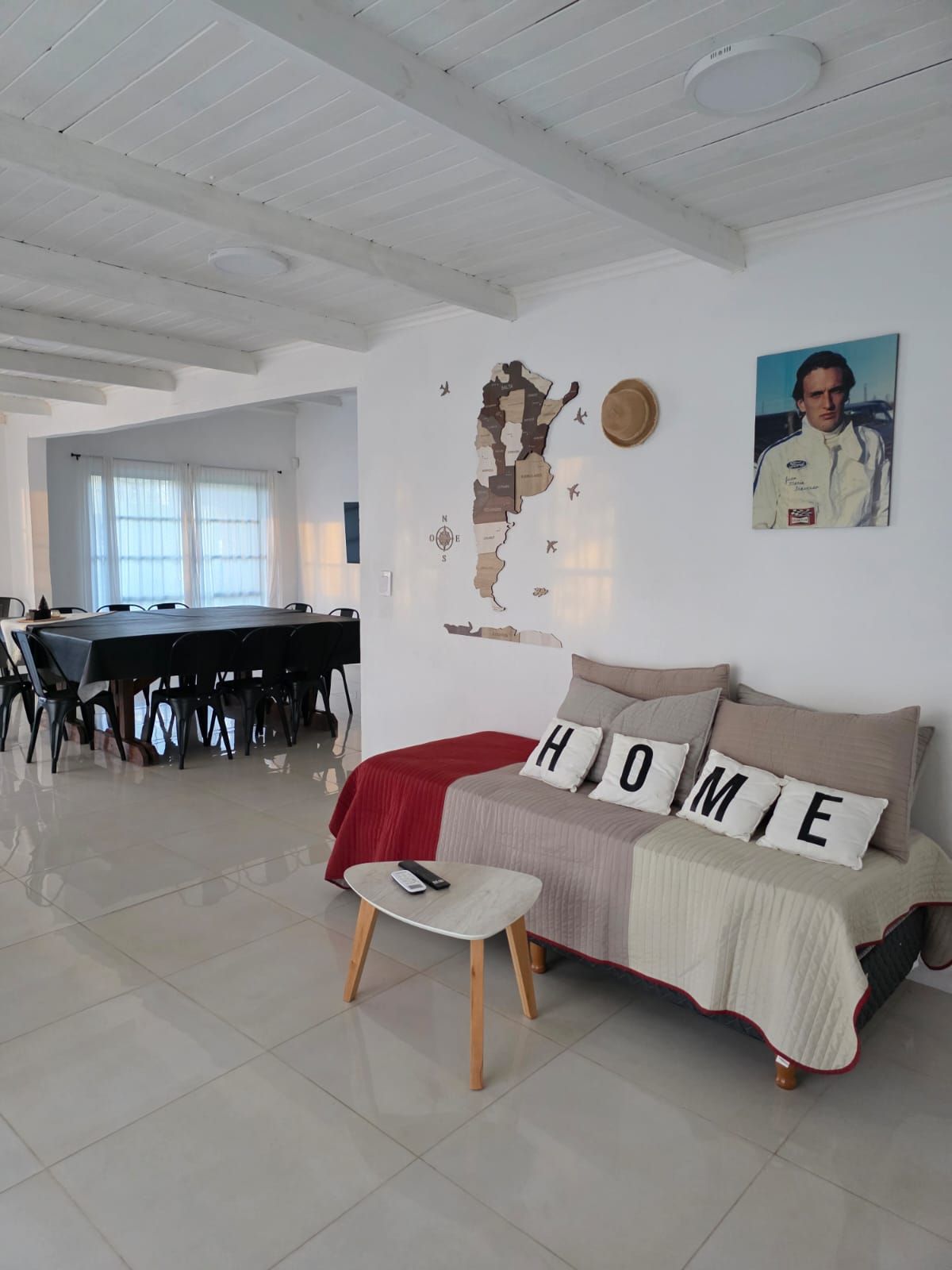 Sala de estar blanca y luminosa con sofá, cojines HOME, mesa pequeña, zona de comedor y cuadros en la pared.