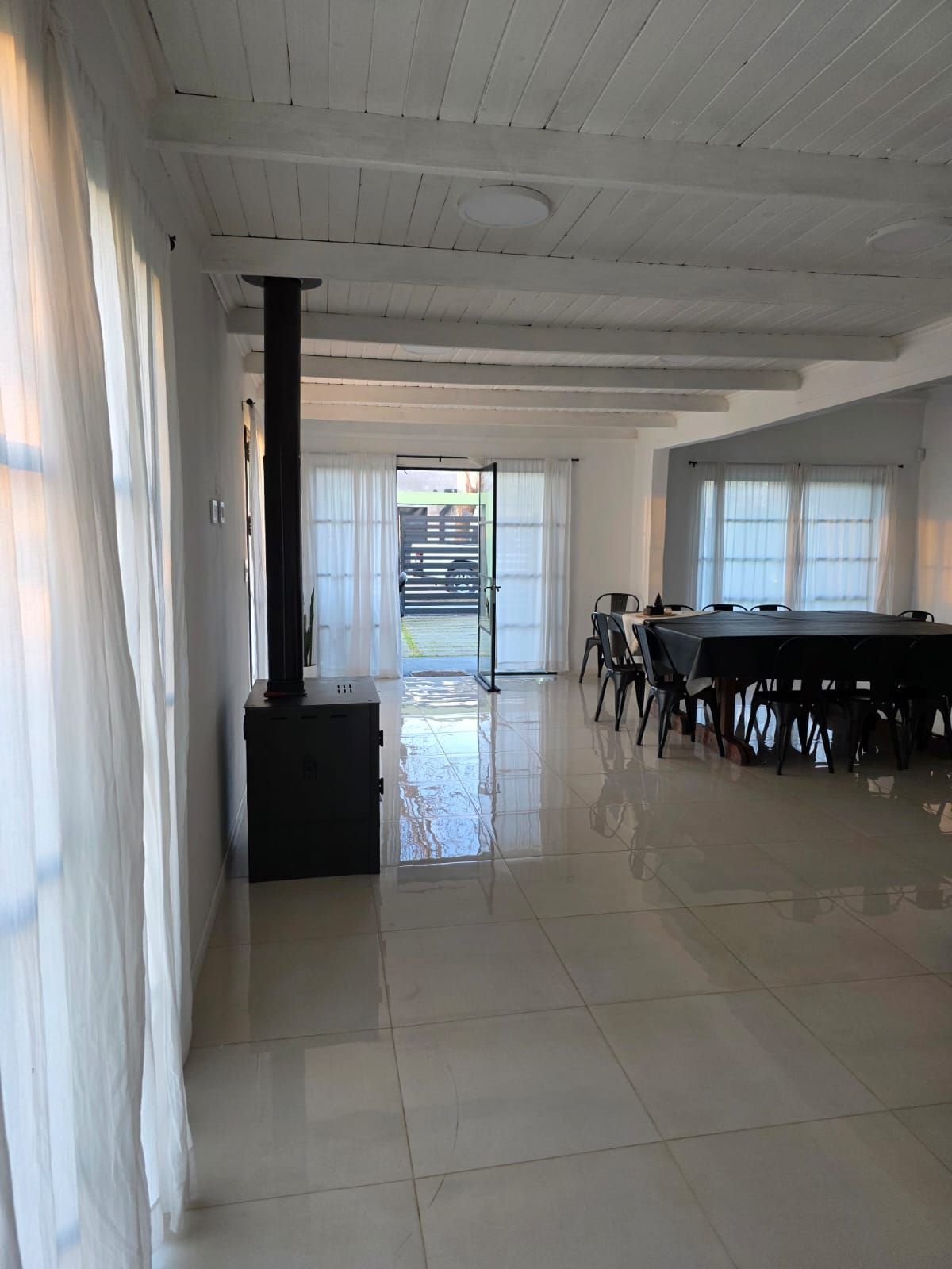 Comedor luminoso con suelo de baldosas, paredes blancas, cortinas, una estufa negra y una mesa larga con sillas.