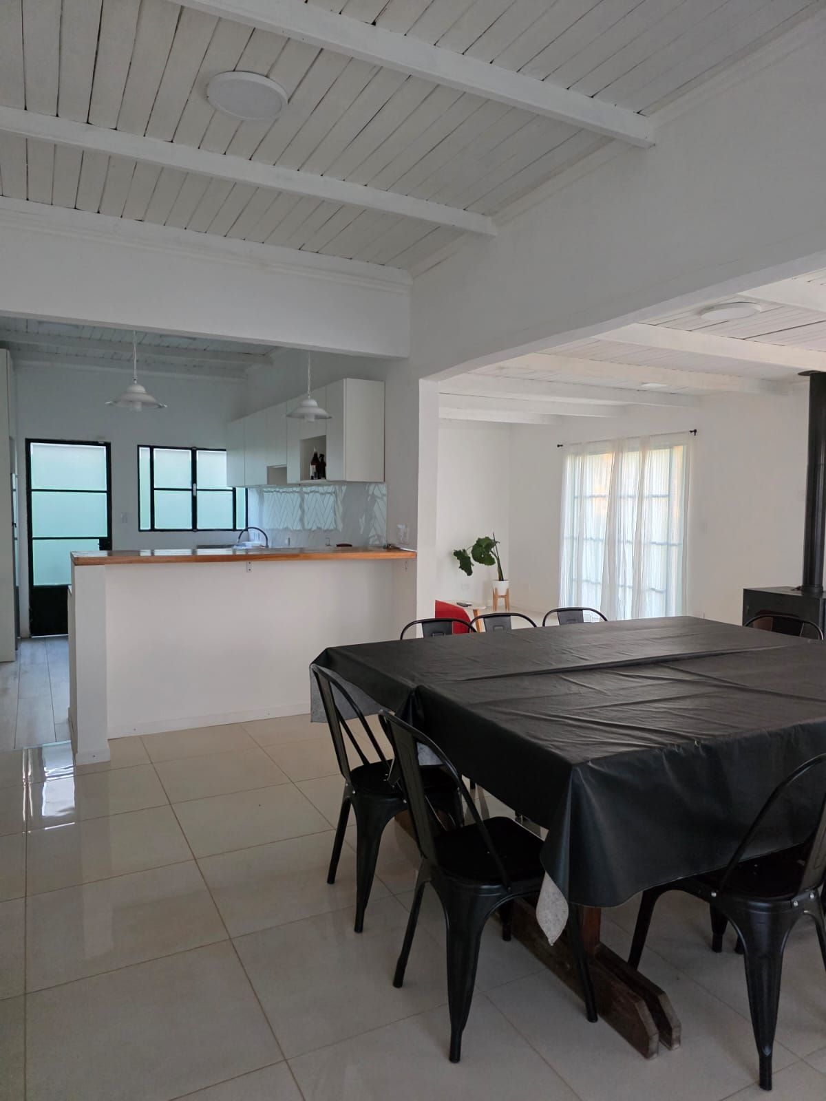 Comedor luminoso con mesa y sillas negras, suelo de baldosas blancas y una encimera de cocina al fondo.