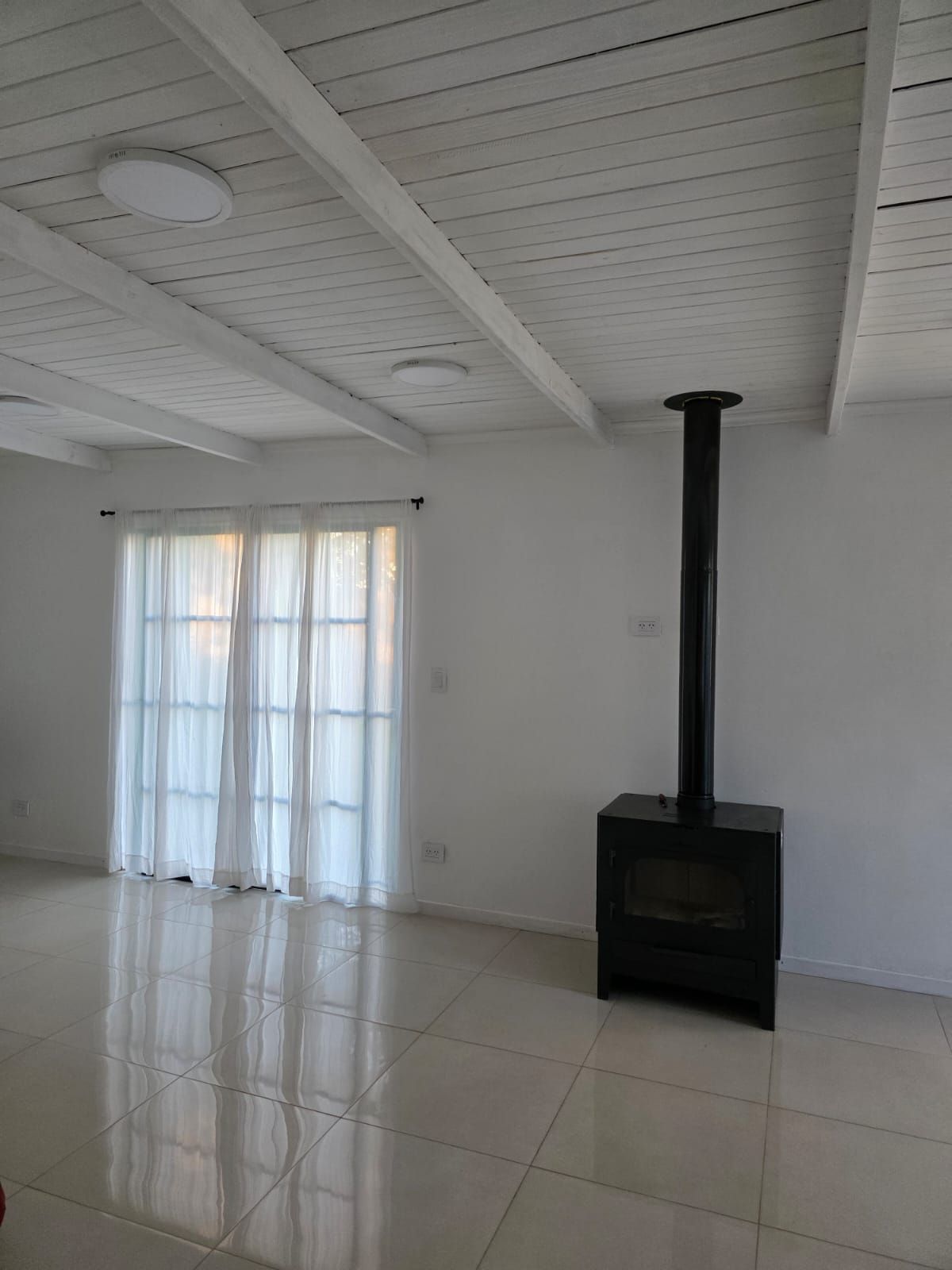 Habitación minimalista blanca con suelo de baldosas, ventana con cortinas transparentes y estufa de leña negra con chimenea.