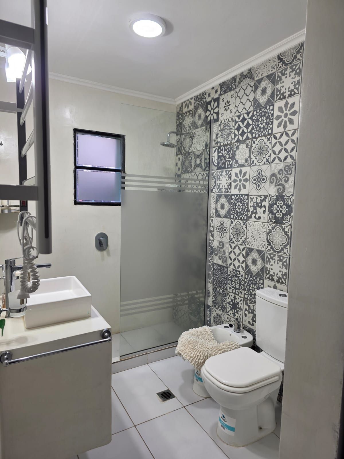 Baño pequeño con azulejos, inodoro, lavabo, ducha de cristal y pared decorativa con estampado.