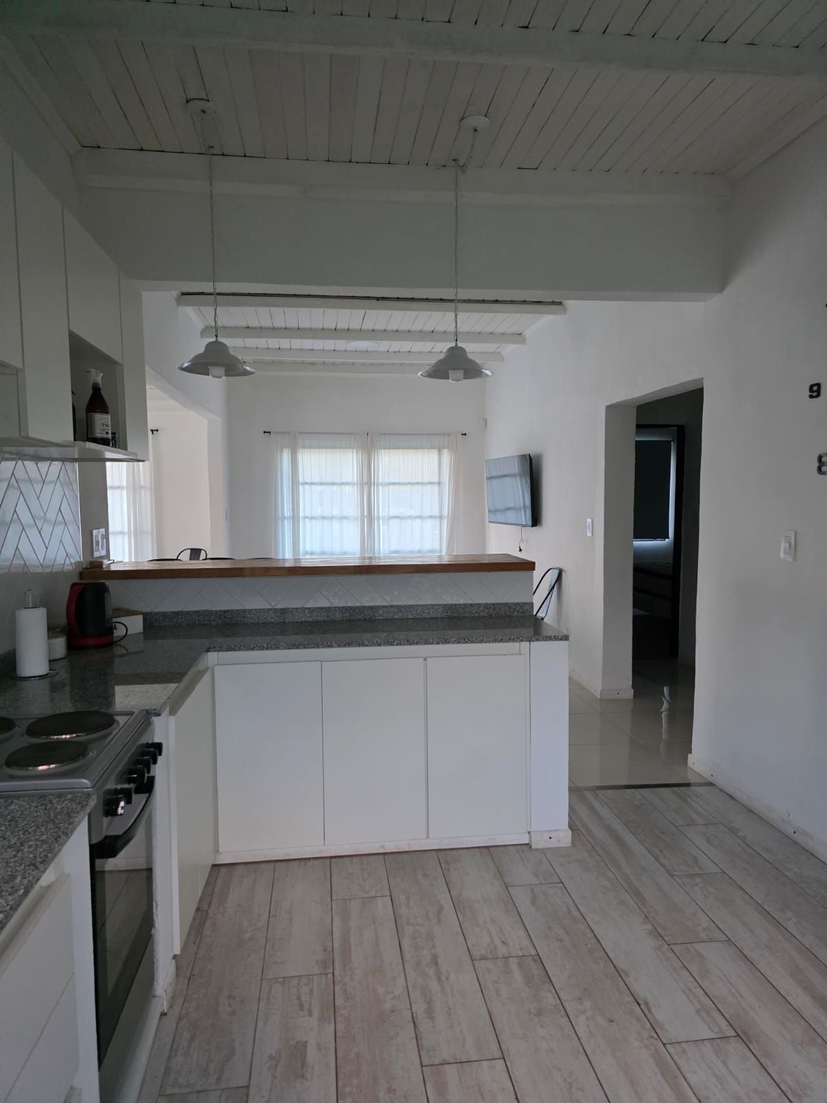 Cocina blanca con isla, suelo de madera, lámparas colgantes y puerta abierta.