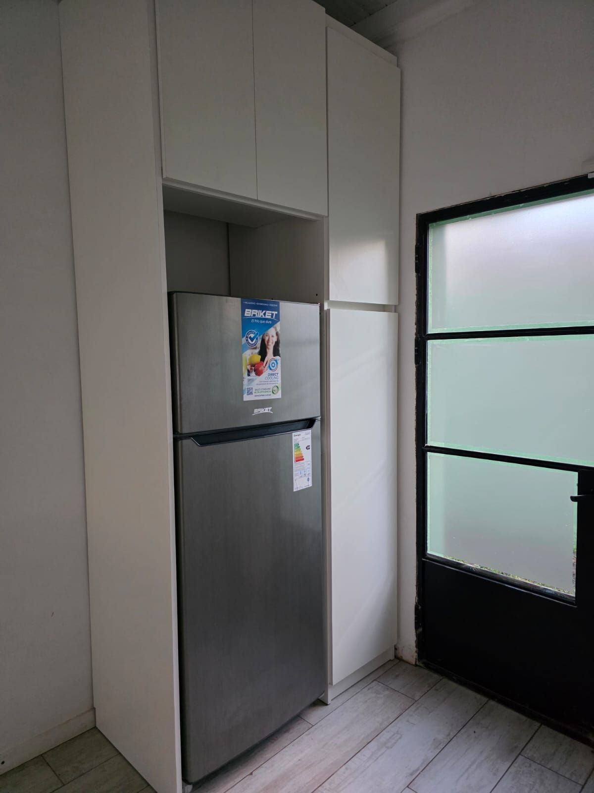 Cocina compacta con un frigorífico plateado junto a una puerta de cristal esmerilado y armarios blancos.