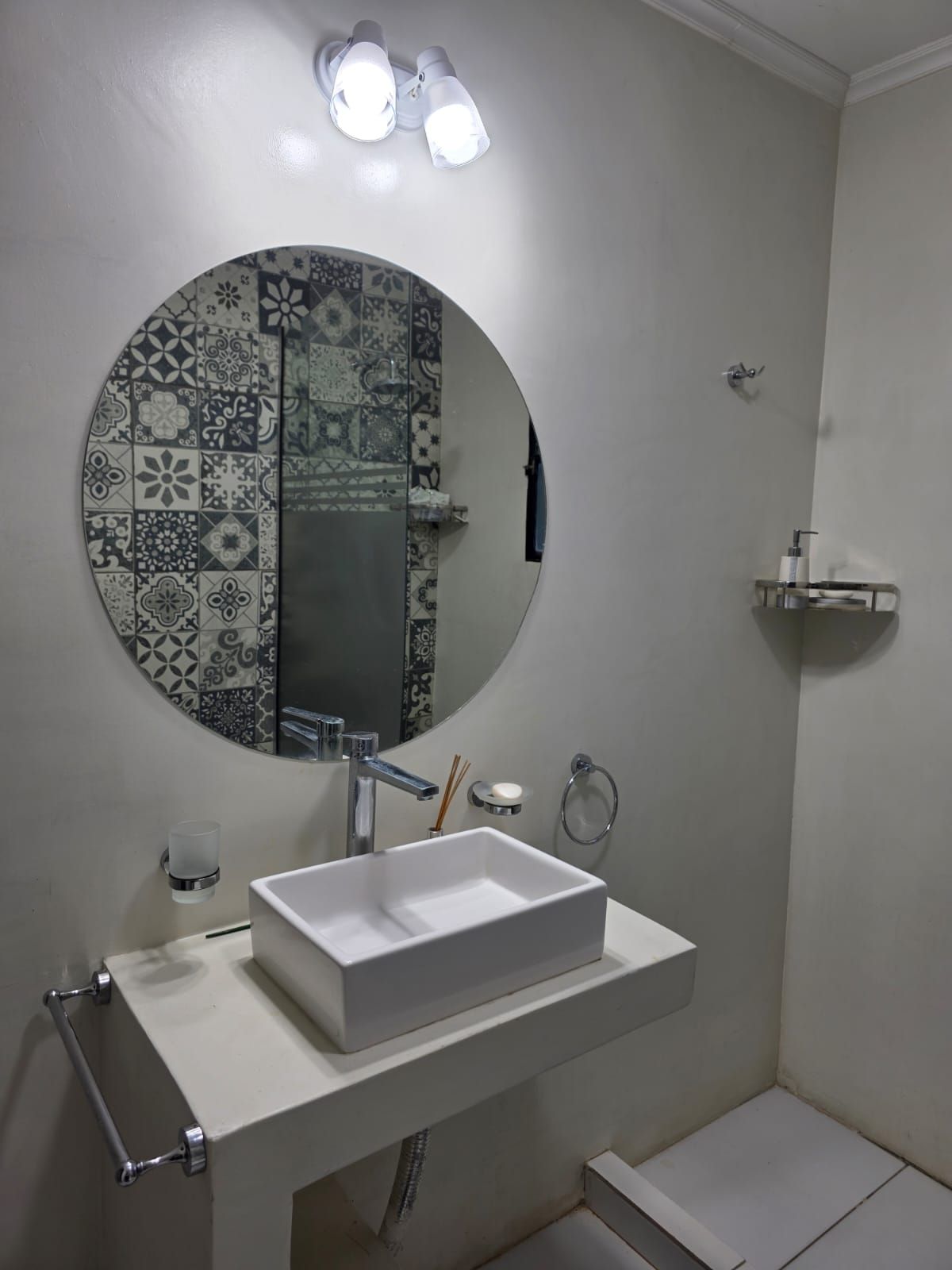 Baño moderno con espejo redondo, lavabo blanco sobre encimera, detalles de azulejos estampados y una pequeña repisa de pared.