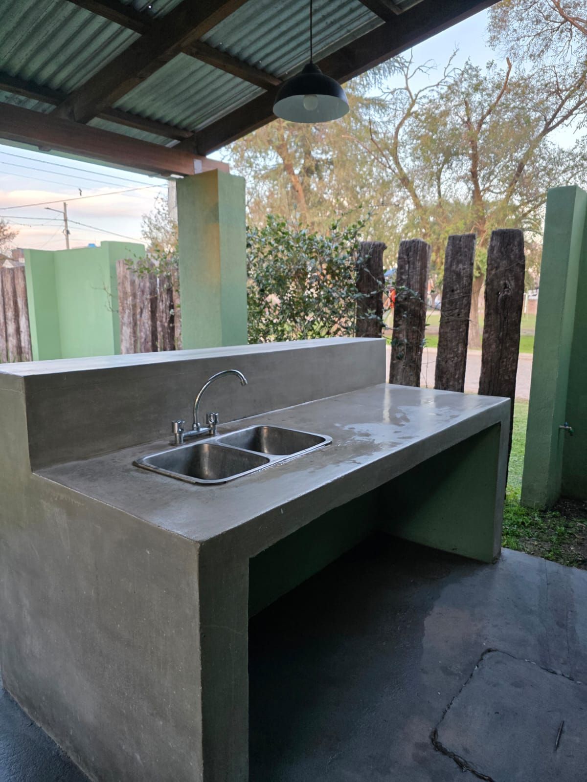 Fregadero y encimera de hormigón para exteriores, situados bajo un patio cubierto, con grifo y dos lavabos.
