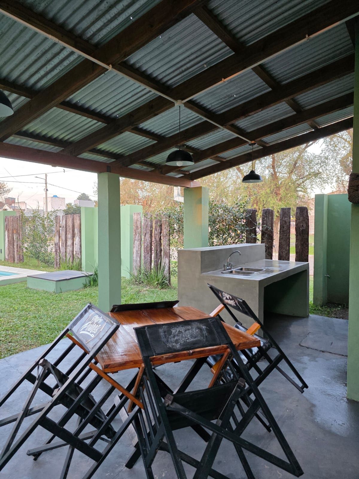 Patio exterior cubierto con sillas plegables negras, una mesa y una parrilla integrada, junto a un jardín con césped.