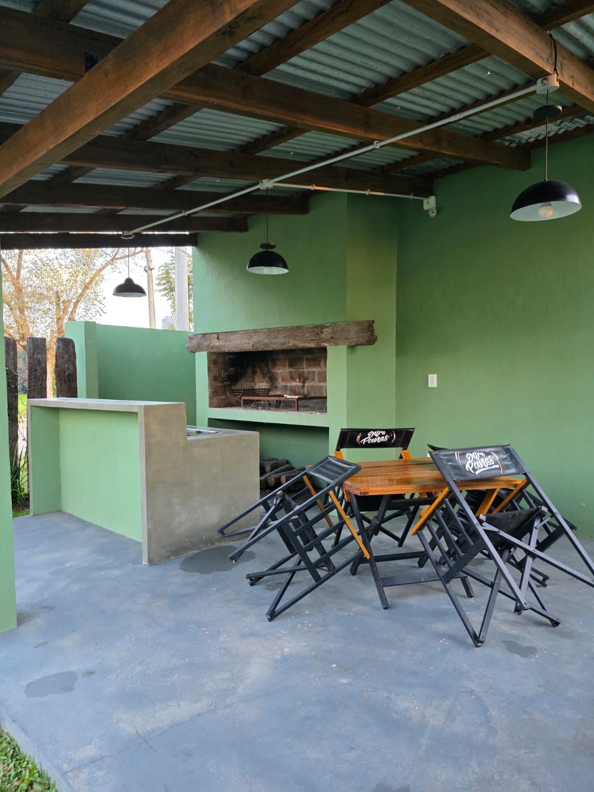 Patio cubierto con paredes verdes, suelo de hormigón, mesa, sillas plegables y parrilla de ladrillo integrada.