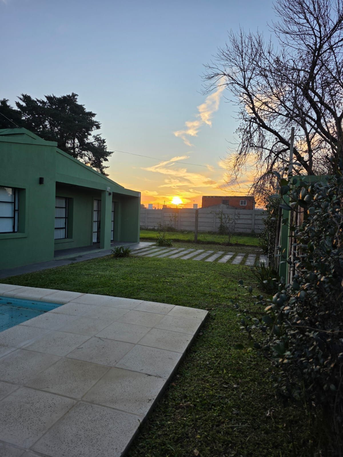 Jardín trasero al atardecer con un invernadero, baldosas de patio, césped y árboles recortados contra la silueta.