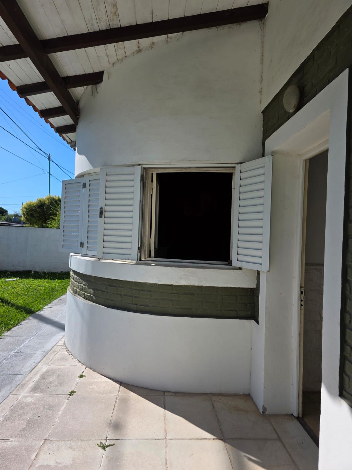 Exterior de una casa blanca con ventana con contraventanas abiertas, pared curva y pasarela con porche sombreado.