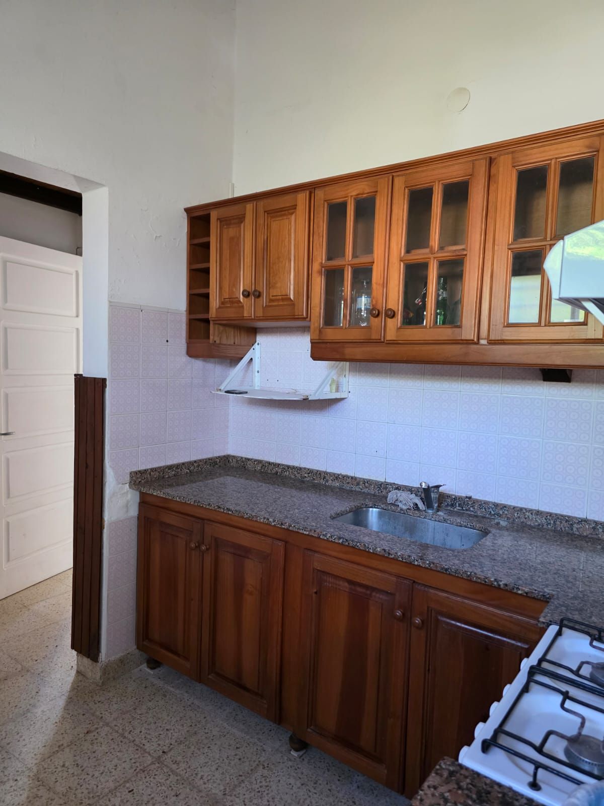 Pequeña cocina con armarios de madera, encimera de granito, fregadero y cocina de gas junto a una pared alicatada.