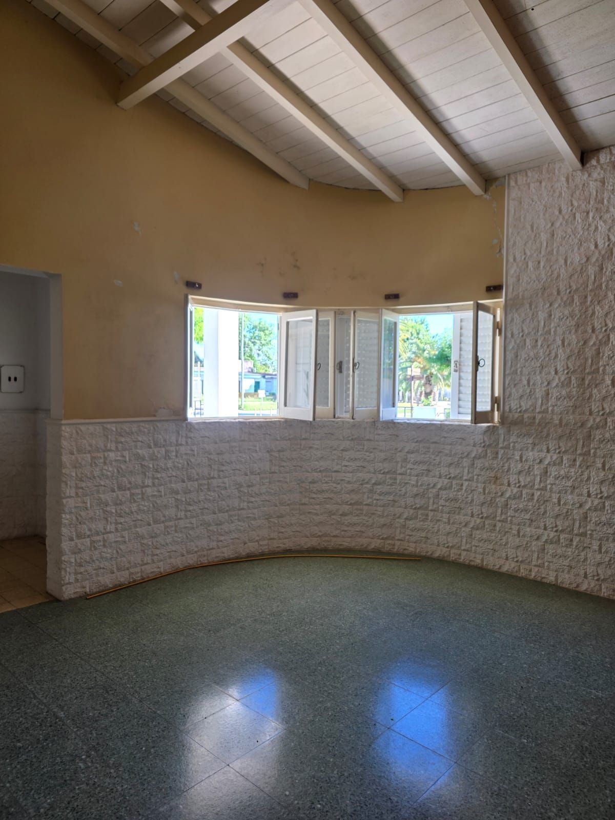 Habitación vacía con media pared de ladrillo blanco, ventanas abiertas y techo abovedado de madera.