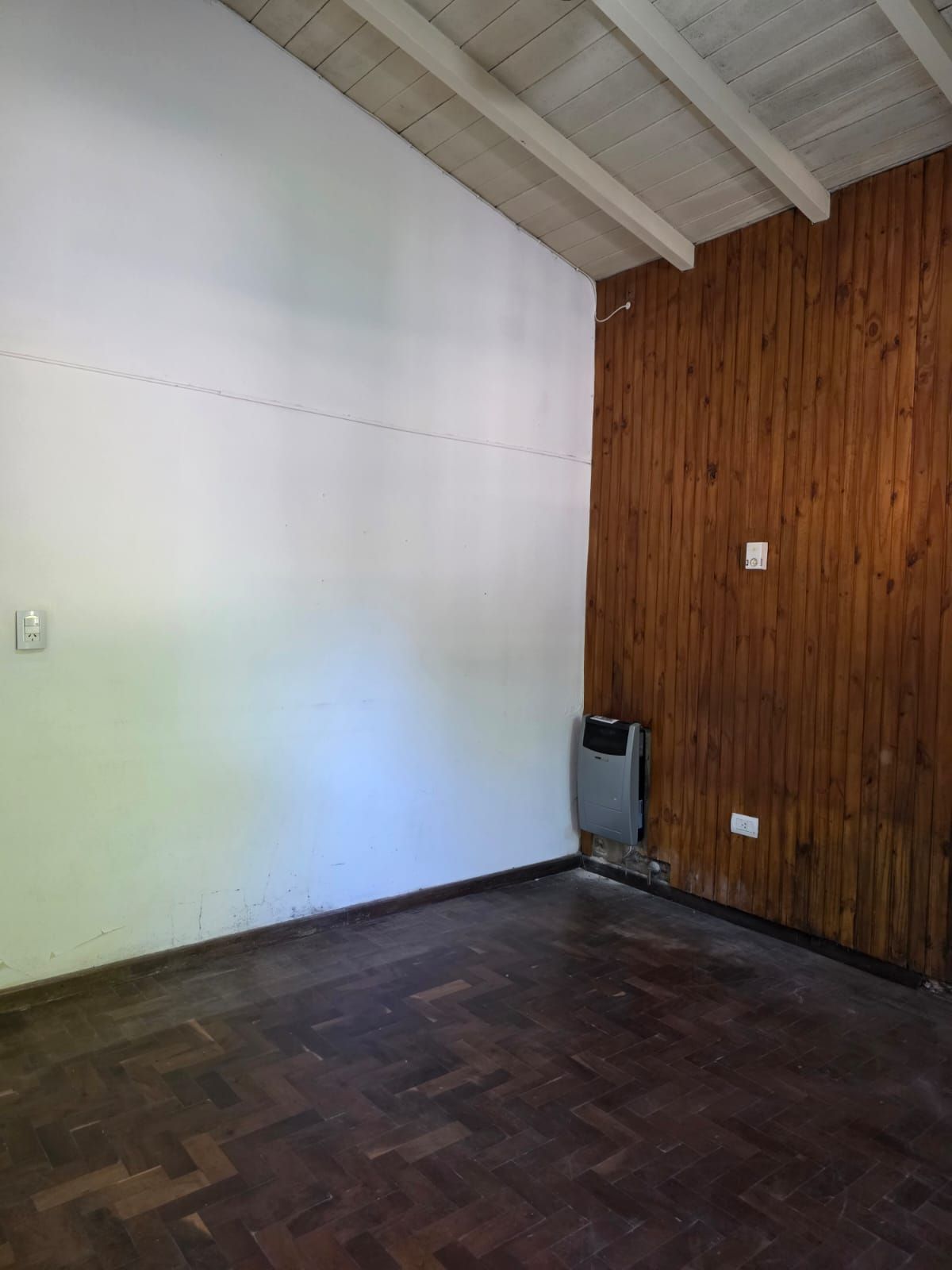 Habitación vacía con paredes blancas y revestidas de madera, suelo oscuro y un pequeño calefactor en la esquina.