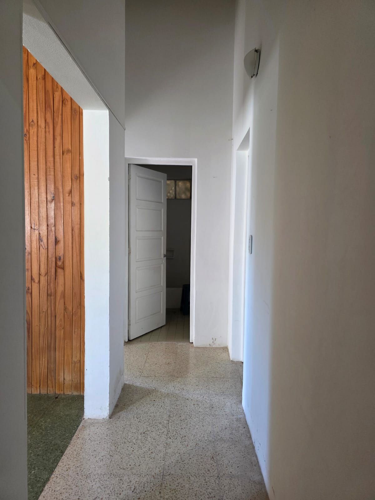 Pasillo estrecho con paredes blancas, suelo moteado, puerta abierta y revestimiento de madera a la izquierda.