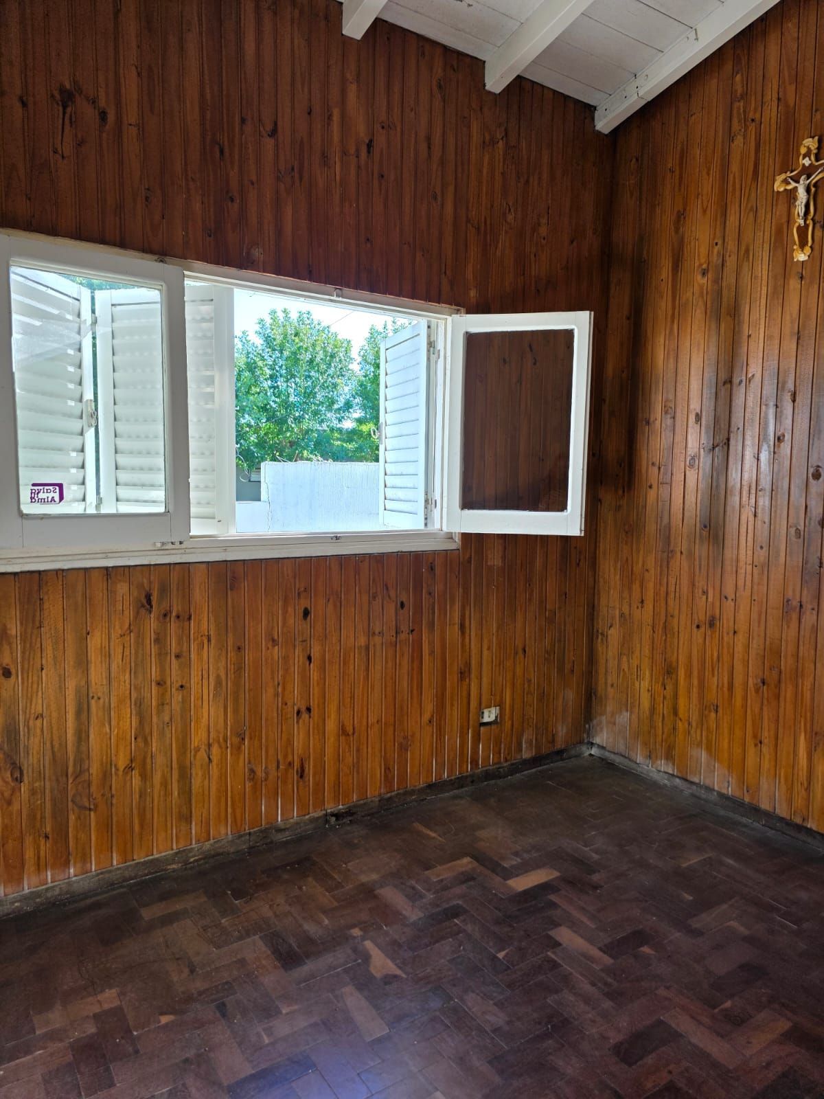 Habitación vacía con paneles de madera, ventana abierta, suelo de parqué oscuro y vigas blancas en el techo.