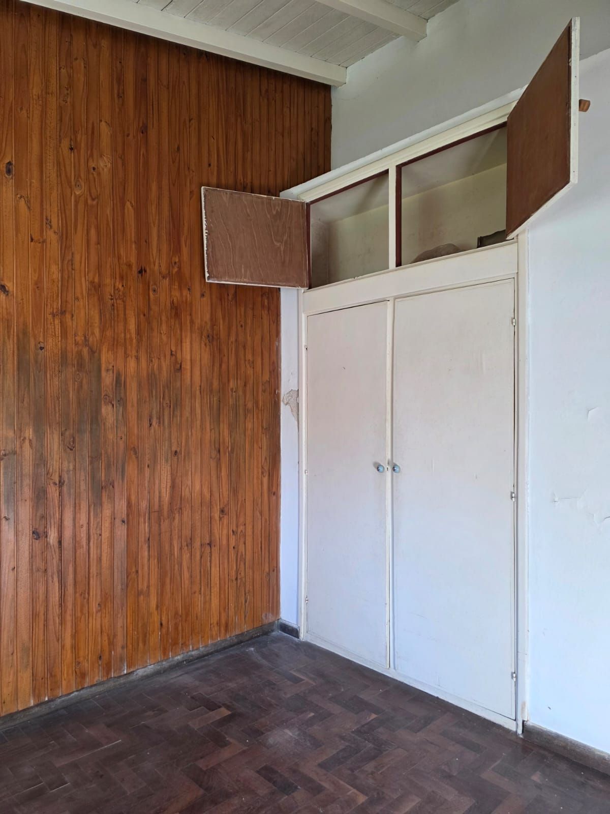 Habitación vacía con pared revestida de madera, puertas de armario blancas y un pequeño armario superior.