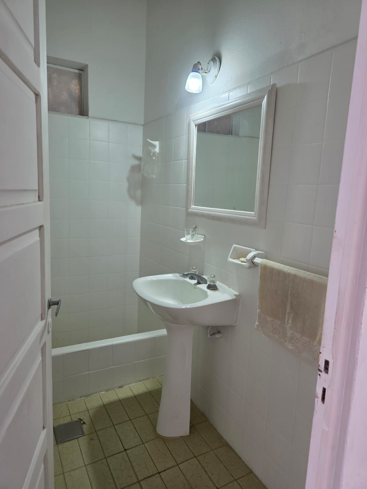 Baño pequeño con azulejos blancos, lavabo con pedestal, espejo, cabina de ducha y toallero.