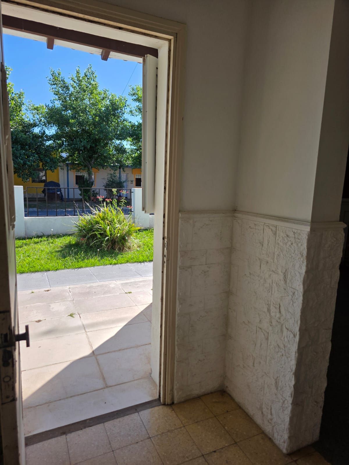 Entrada abierta a una acera soleada y árboles, con paredes interiores y suelo de baldosas en primer plano.