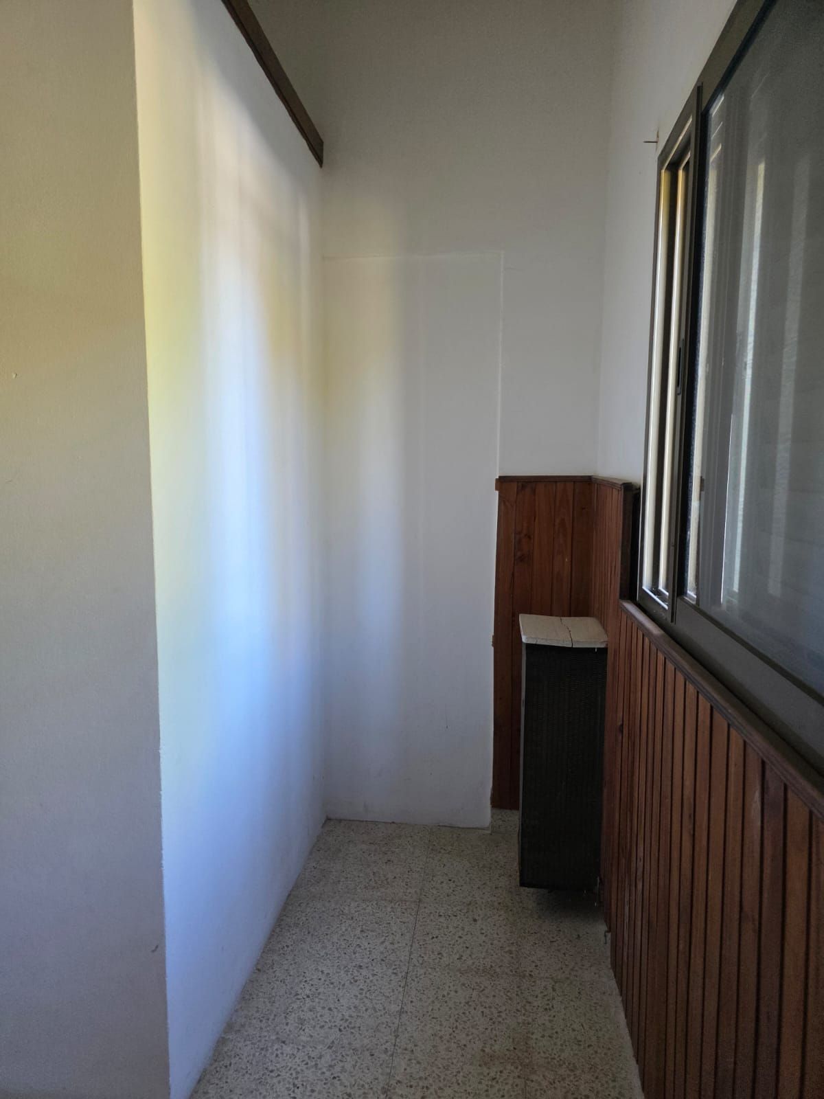 Balcón estrecho con paredes blancas, suelo de baldosas y barandilla de madera junto a las ventanas.